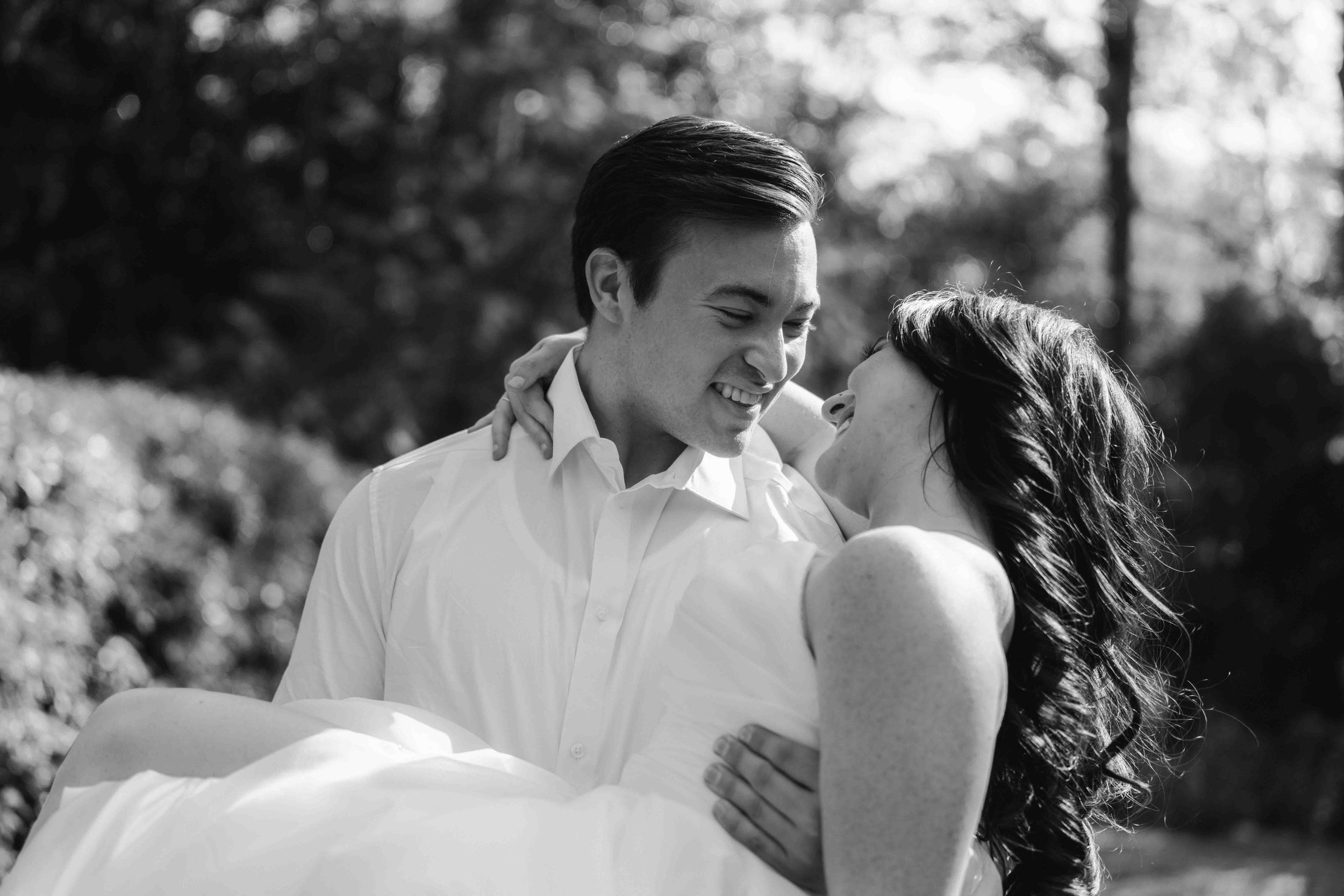 A groom lifts a bride into his arms while both smile toward each other, surrounded by trees and filtered light, observed by a Fragmented Memories prenup photographer.