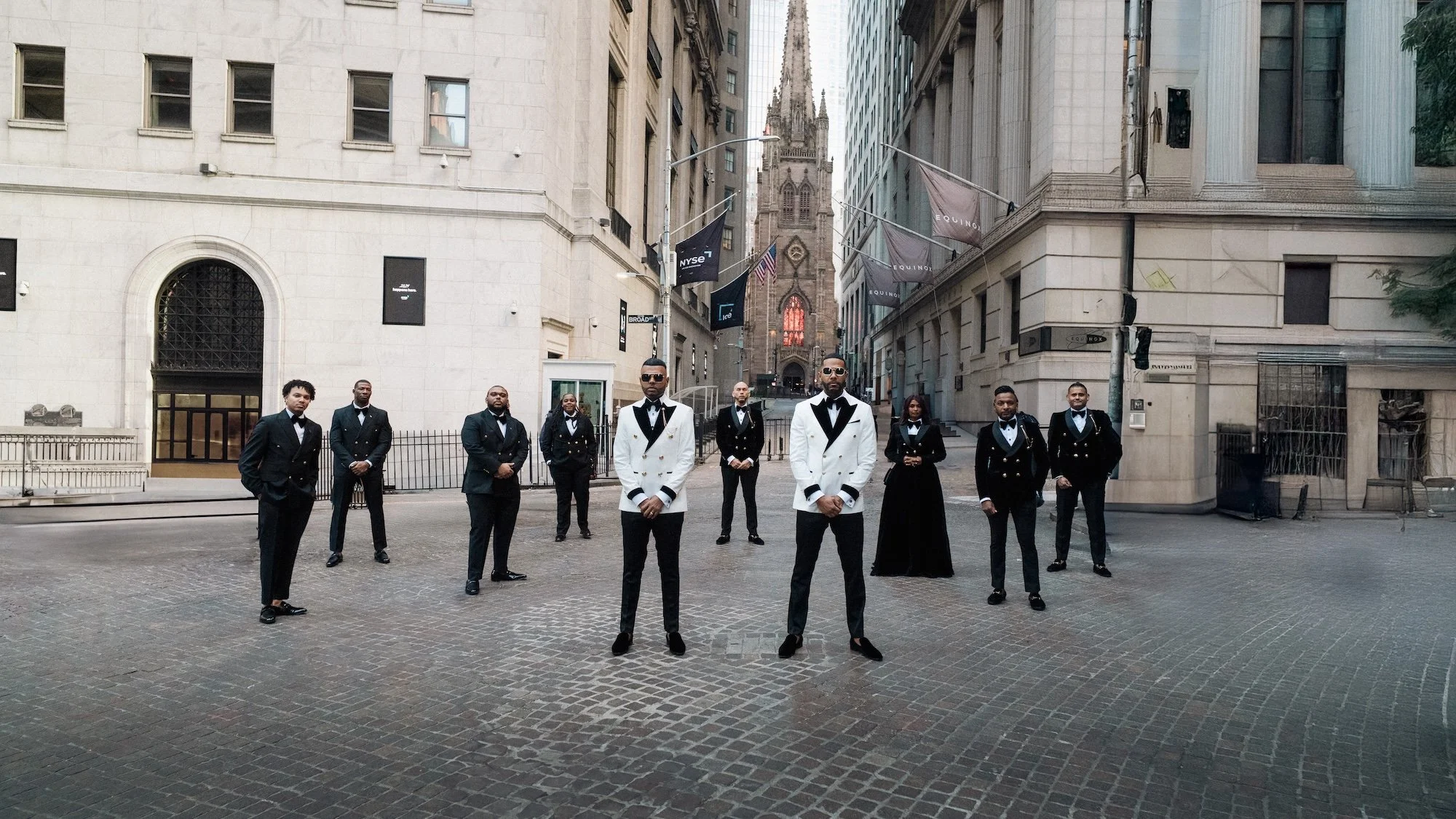 The grooms stand with their wedding party in tuxedos on cobblestone streets near Trinity Church.
