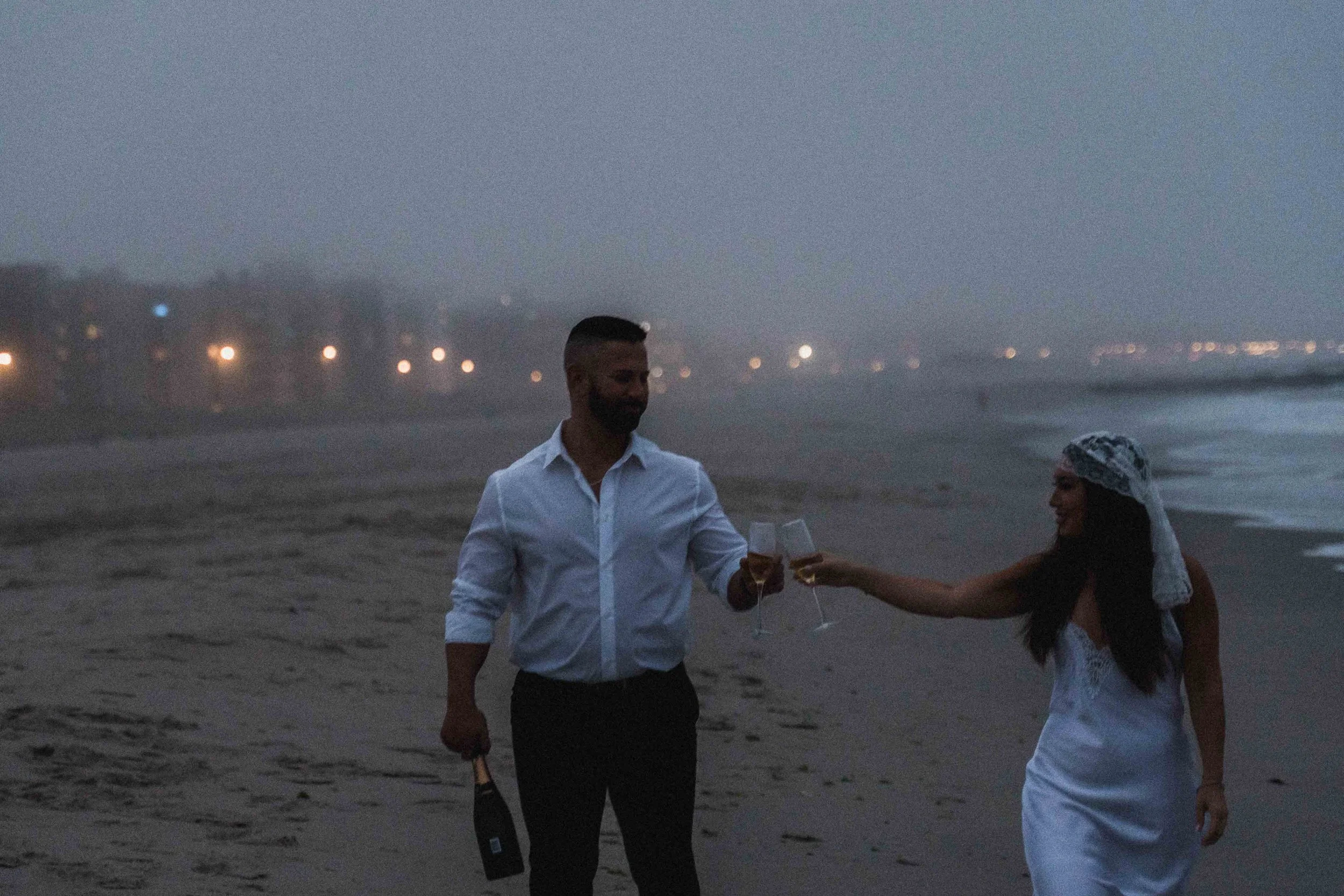 A man in a rolled shirt stands on wet sand holding a bottle as a woman reaches toward him, framed by dusk haze from a Fragmented Memories prenup photographer.

