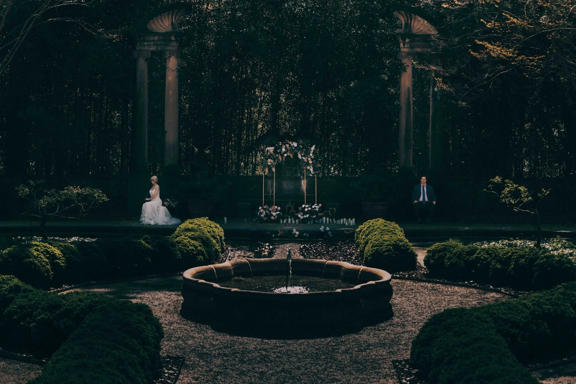 cinematic wide shot of bride and groom framed by a garden fountain and architectural columns.