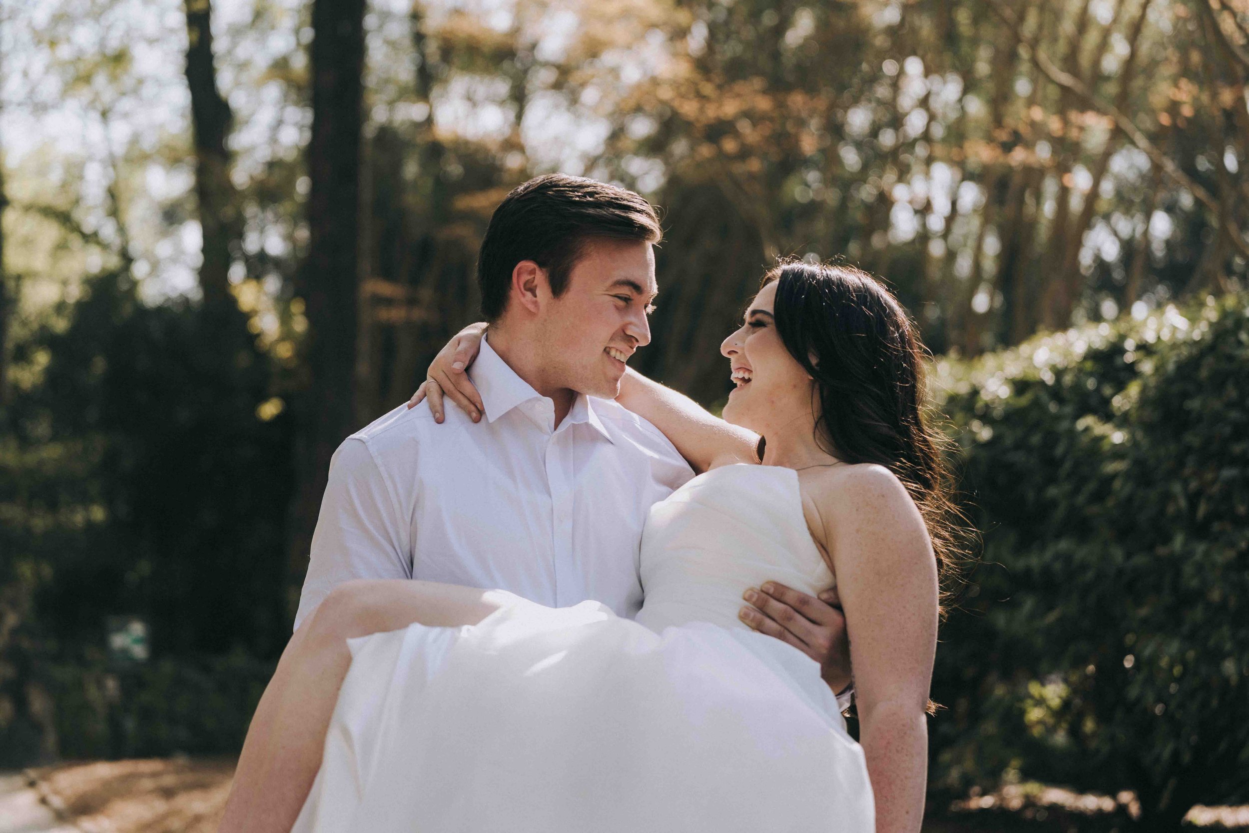 A man carries a woman in a white dress through a wooded path, watched quietly by a Fragmented Memories prenup photographer.