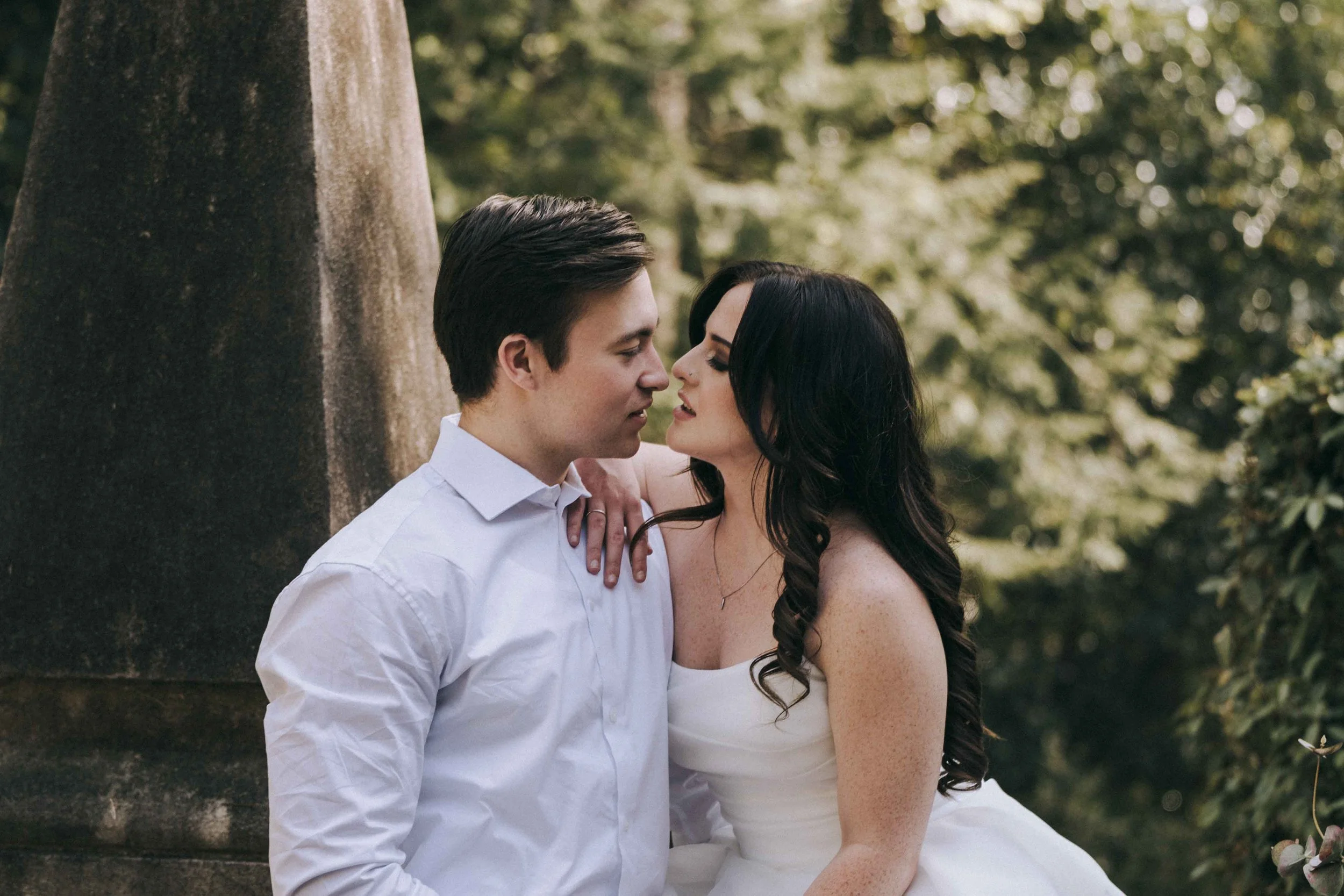 A couple stands beneath trees, foreheads nearly touching, framed by filtered light and a calm, grounded posture.