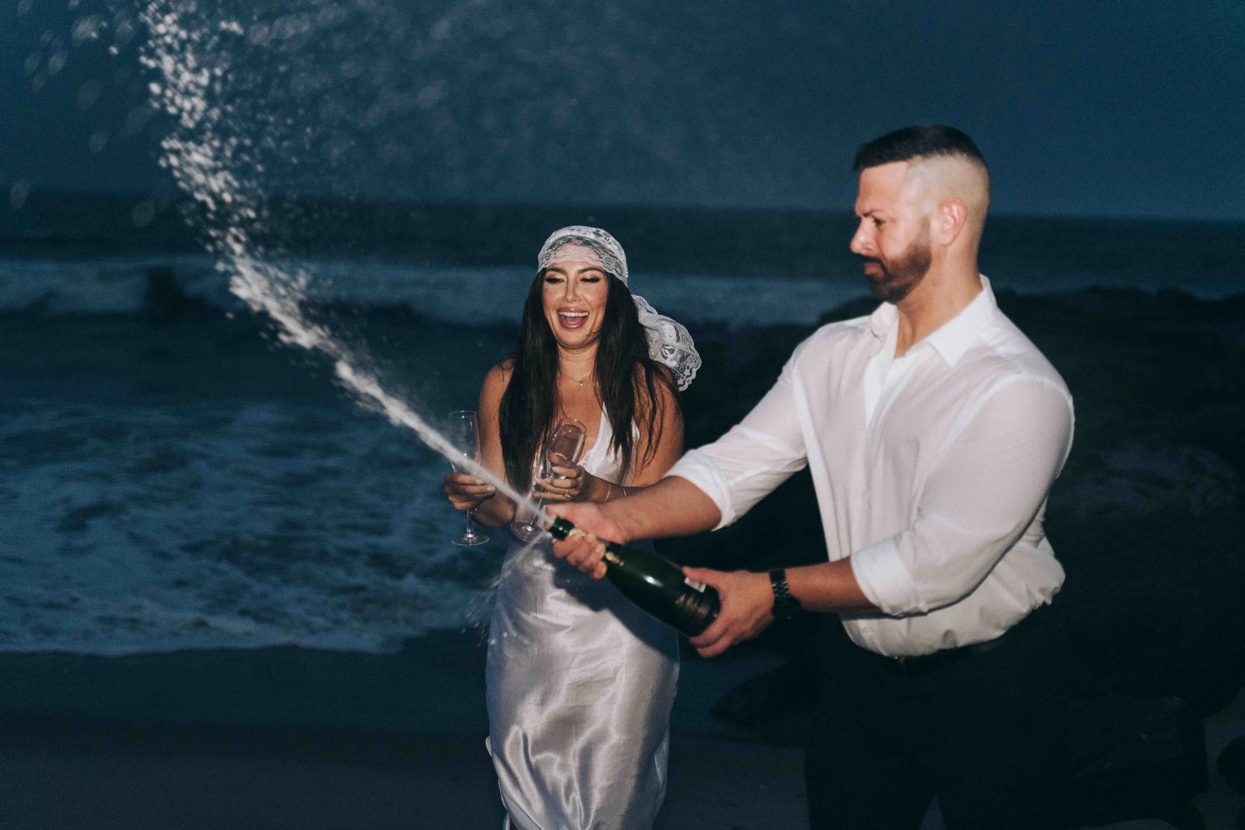 A couple celebrates at night as champagne sprays upward beside the ocean, framed with the energy of a cinematic couples film moment.