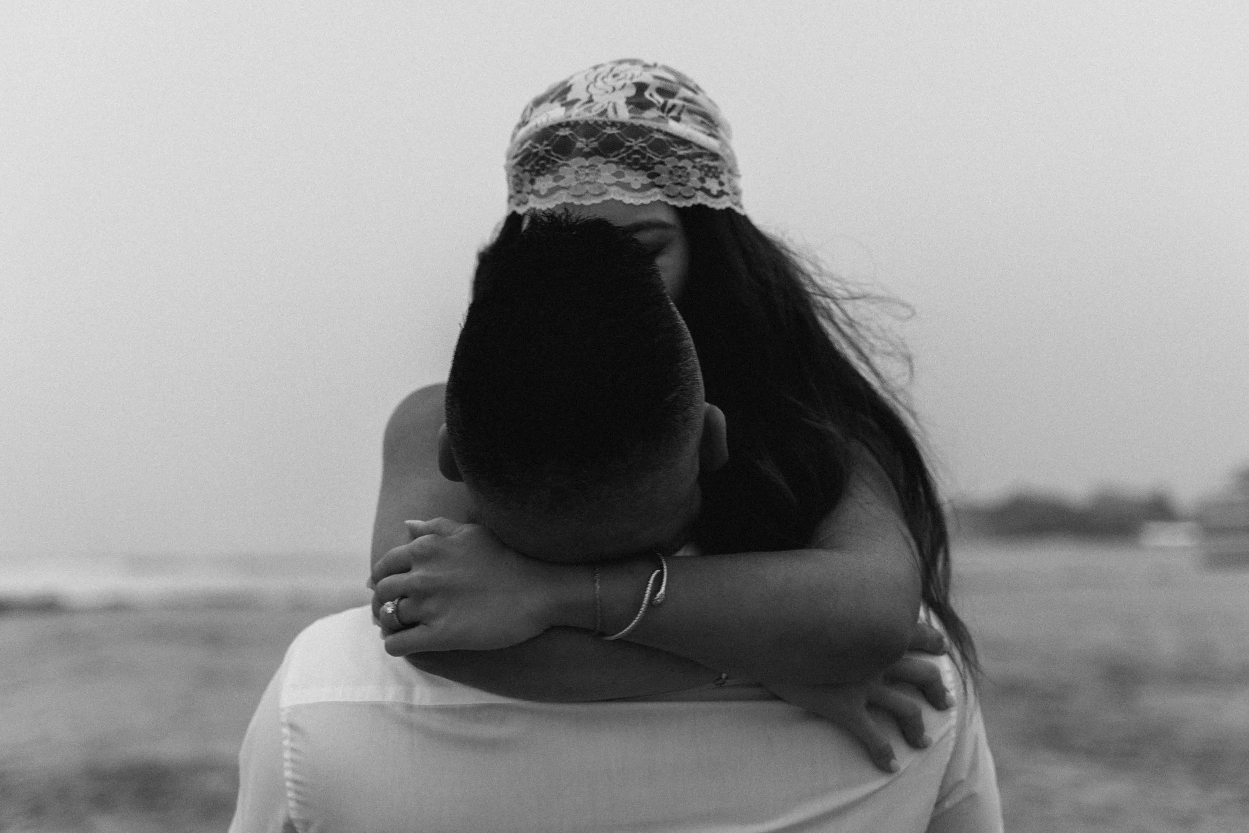 Two figures embrace tightly near the shoreline, bodies pressed together as wind and water move behind them.
