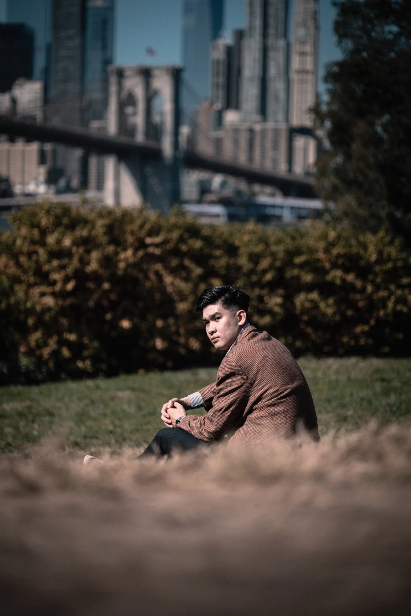 A man sits on the ground in casual attire, framed against trees and architectural lines, inspired by editorial portrait photographer