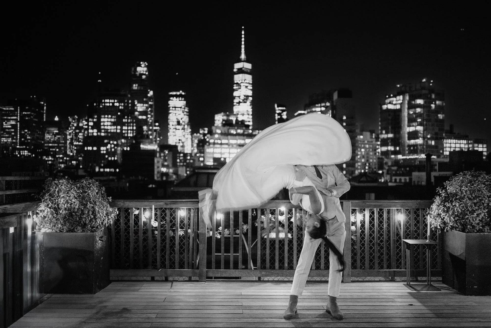 A couple dances on a rooftop at night, city lights glowing behind them as fabric lifts in the wind