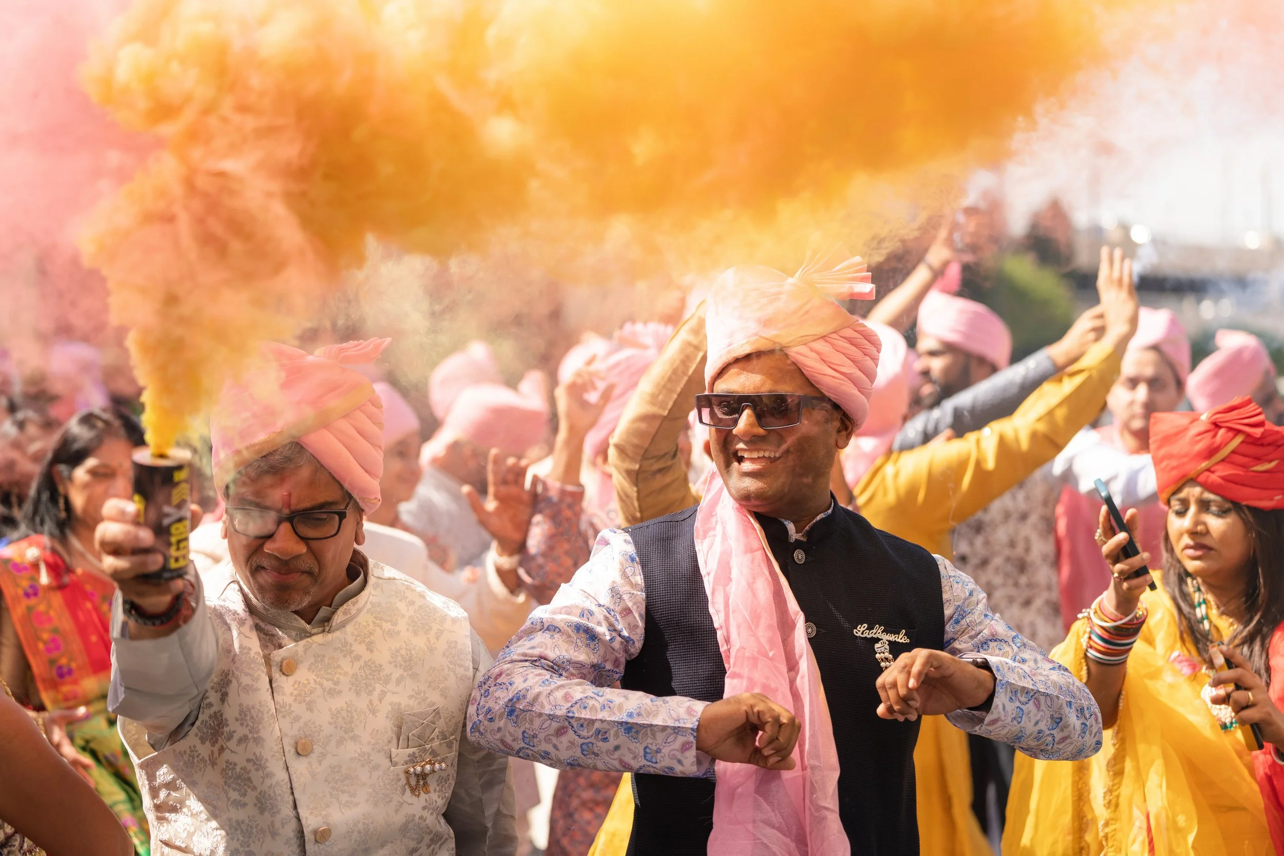 Guests in pink turbans celebrating with arms raised in the midst of colorful smoke, inspired by India weddings