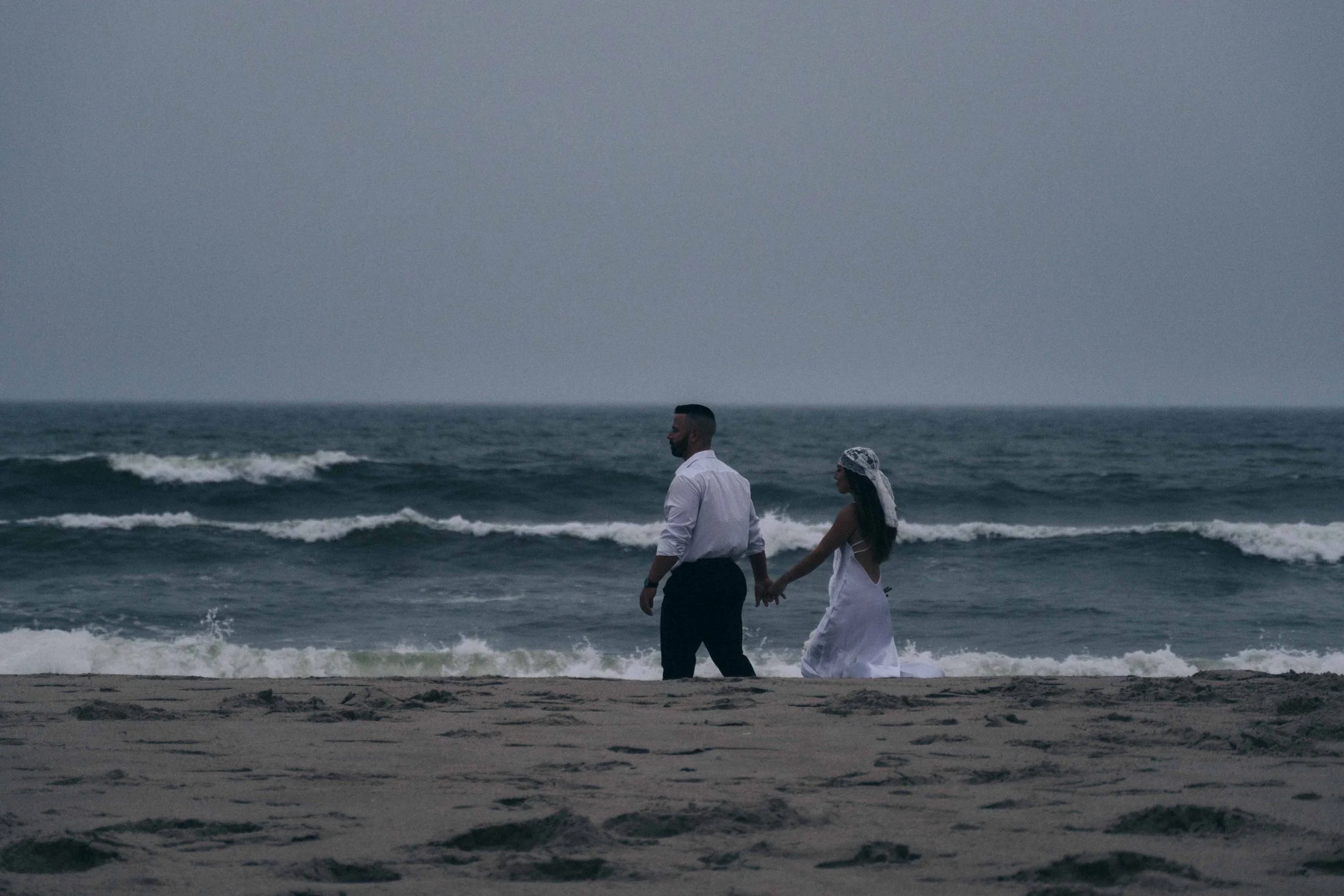 A couple holds each other at the water’s edge as waves break against their legs, framed wide like a cinematic prenup film still.