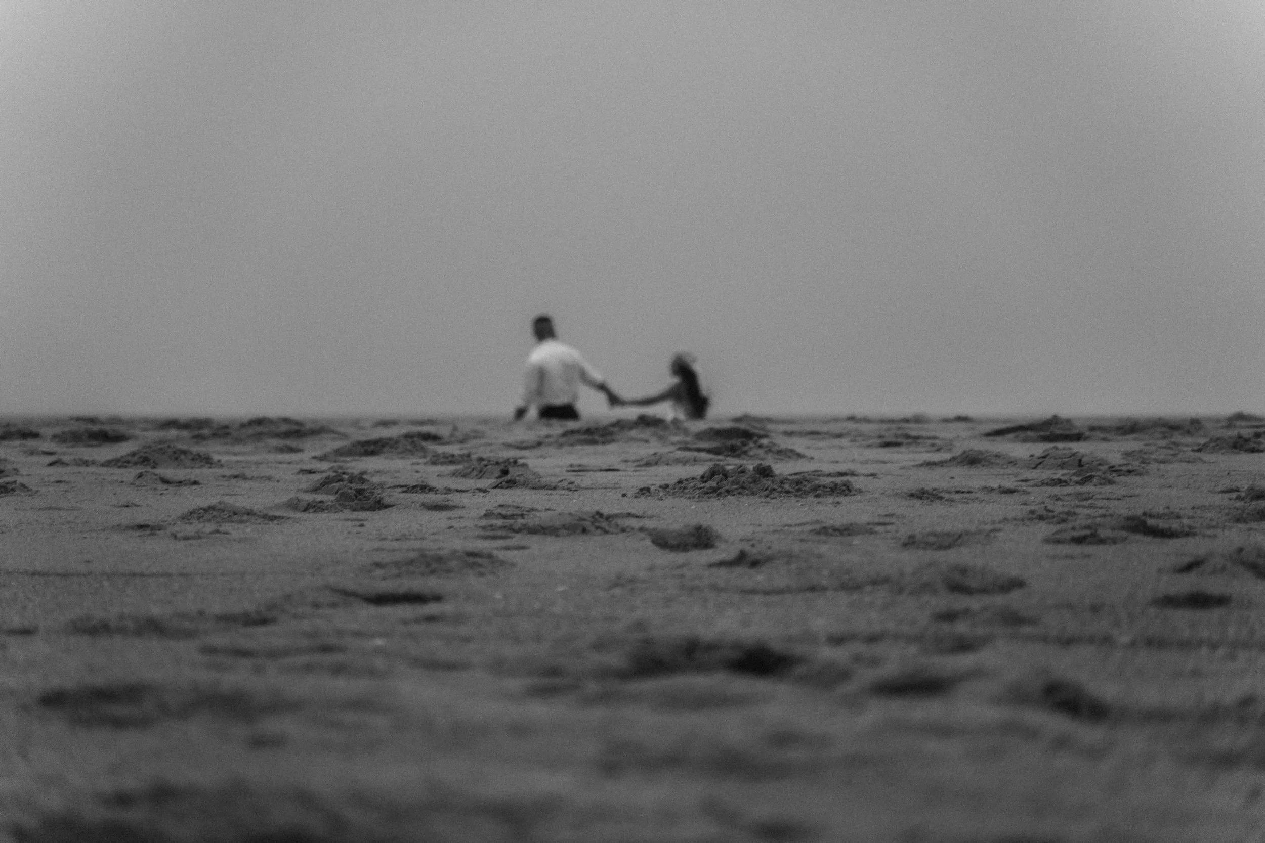 A distant couple holds hands while walking across textured sand, observed quietly without keywords as the frame emphasizes separation and space.