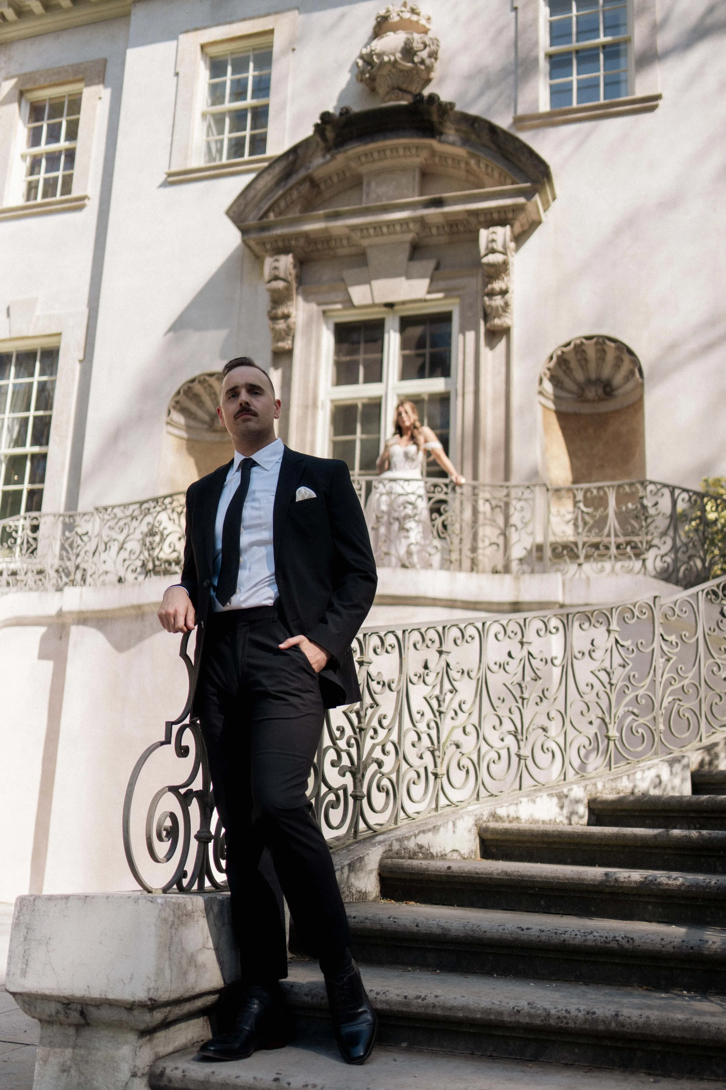 A groom leaning against an ornate railing, his suit sharp in the light, his gaze reflective.