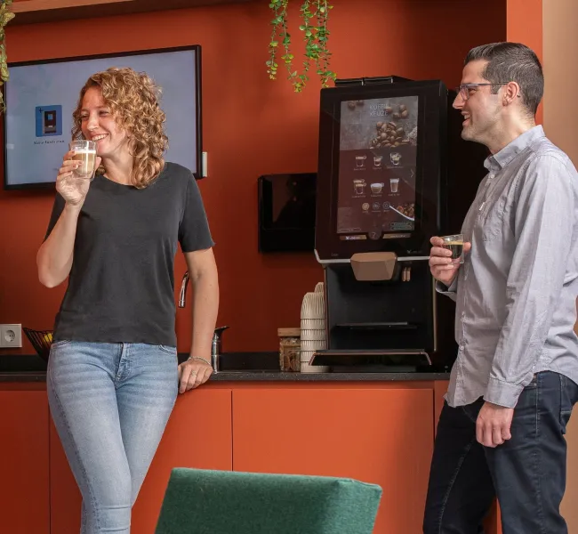 Two people enjoying coffee beside a bean-to-cup coffee machine in an office break area.
