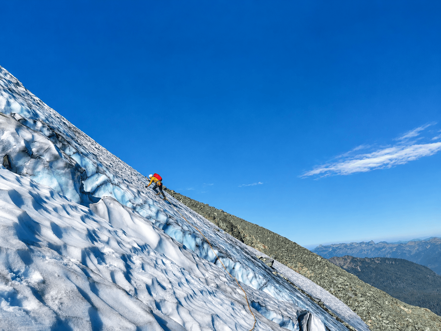 A climber in a yellow jacket and red backpack is leading up a steep snow and ice slope with crevasses nearby on the North Face of Mount Shuksan.
