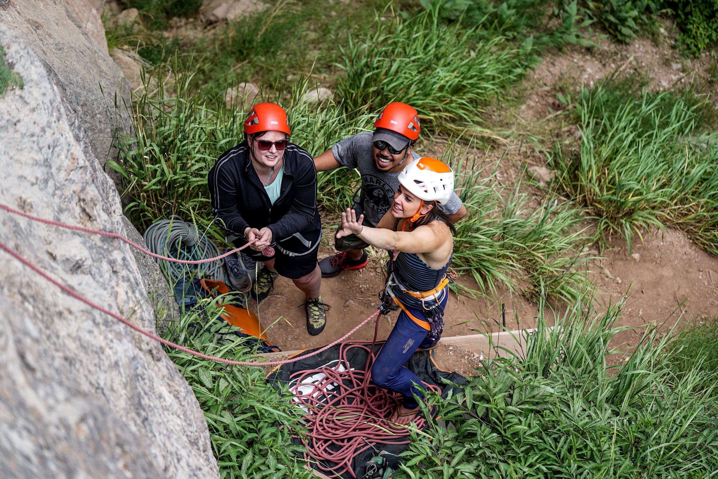 Two climbers, a man and woman in red helmets look up at the cliff as the guide, a woman in a white helmet is pointing and instructing.