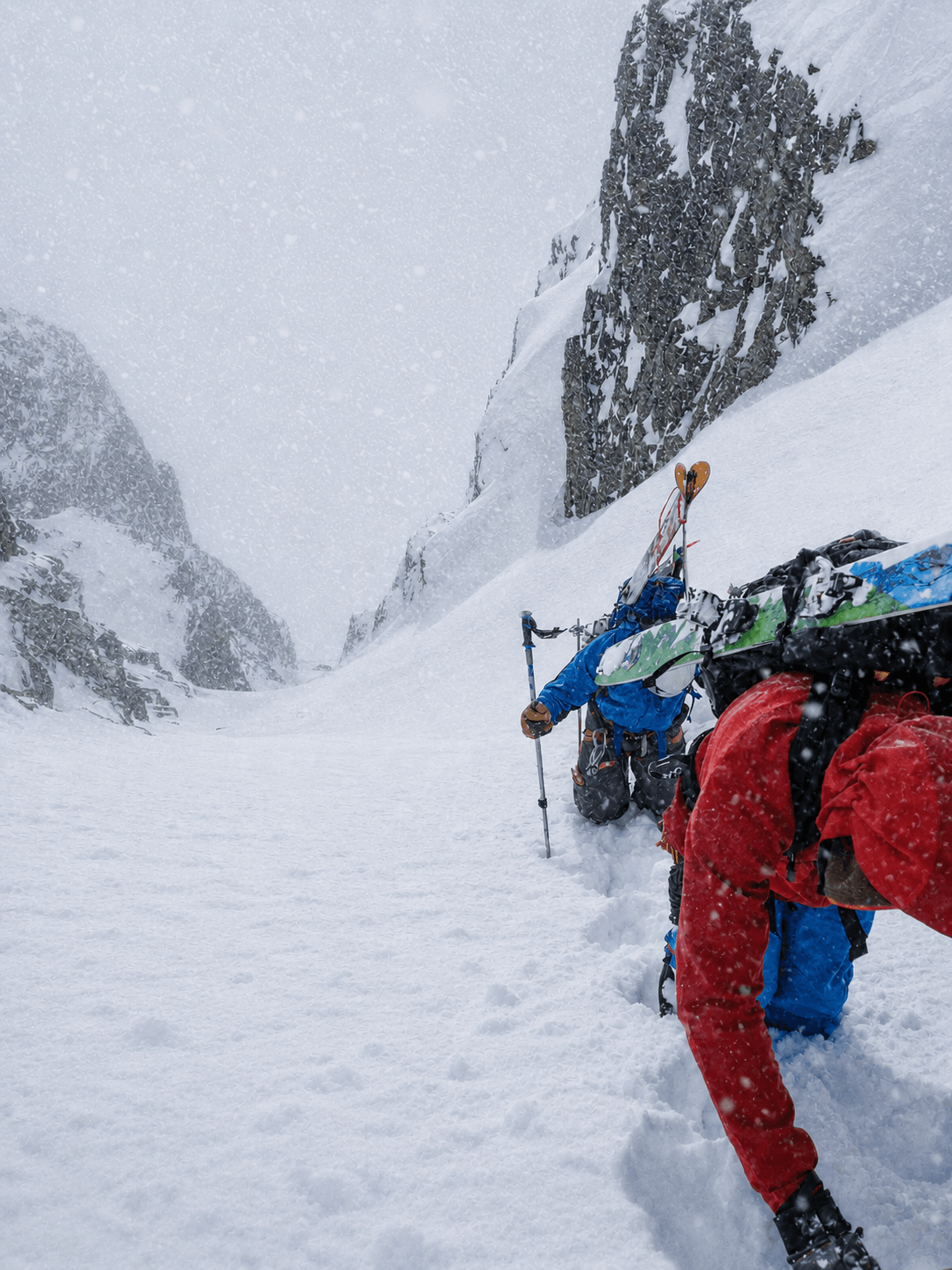 Skiers climb up a steep couloir with their skis on their back.