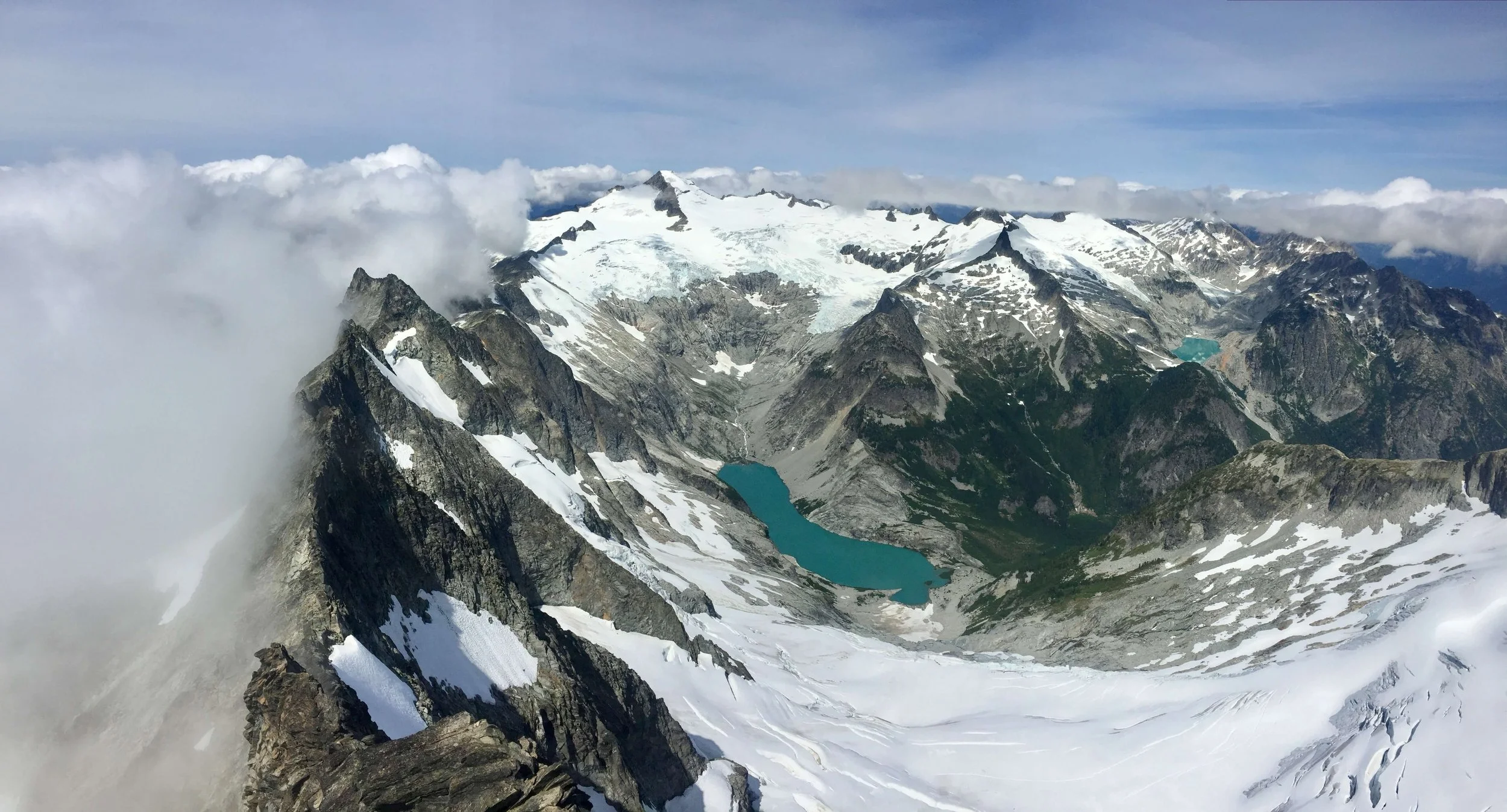 The West Ridge of Forbidden Peak descends into thick clouds with the large glaciers of Eldorado Peak in the background.