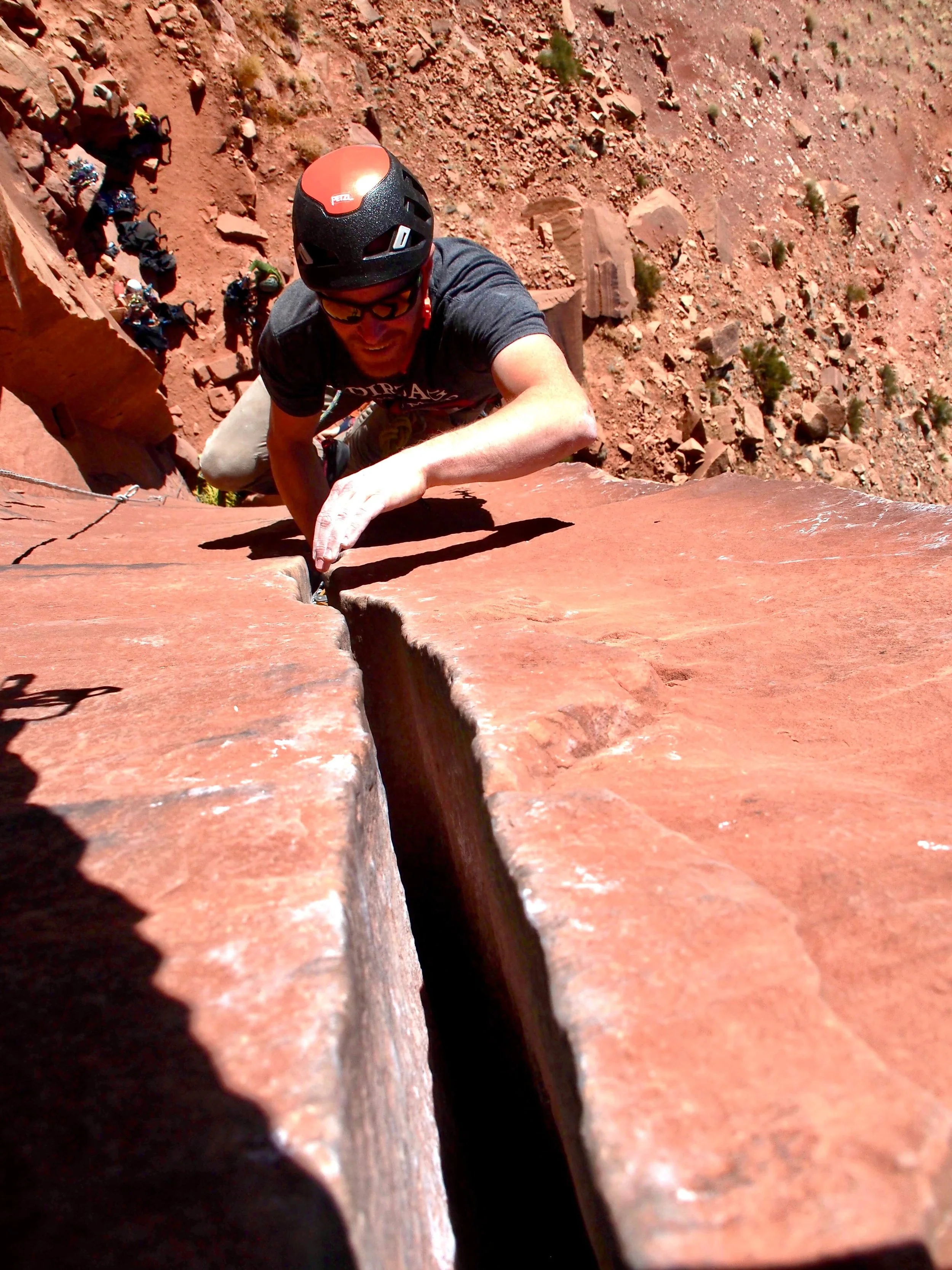 A guide in a black helmet is leading a crack climbing route in Moab, Utah. He is reaching up to jam his hands in the sandstone crack.