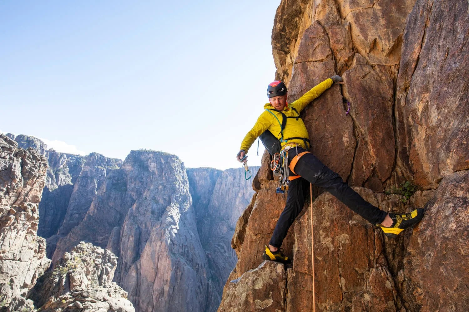 A rock climbing guide in a yellow jacket is leading up a corner route in the Black Canyon. He is reaching down to his harness to place protection.