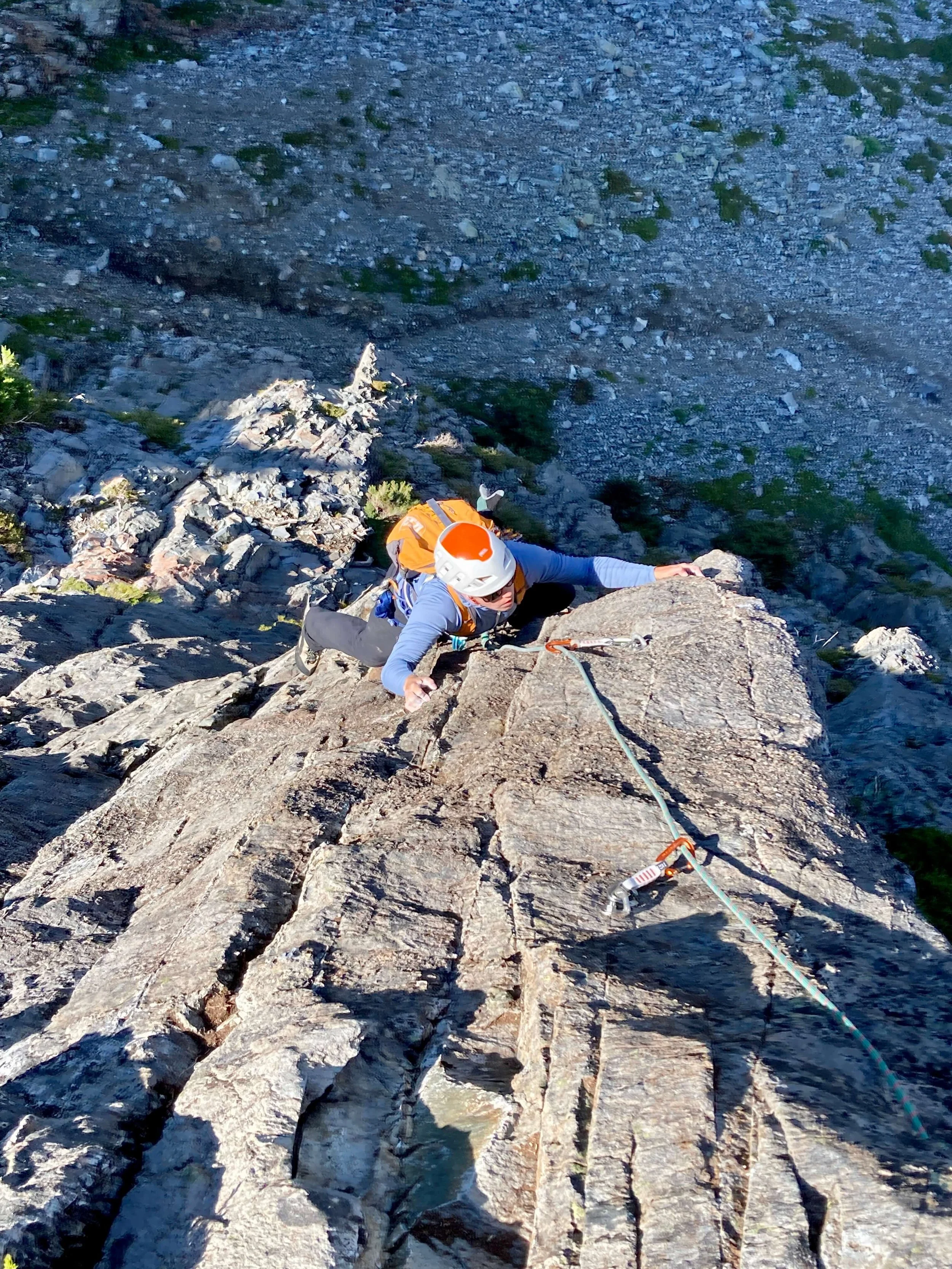 A rock climber in a white and orange helmet with an orange backpack is reaching for a handhold on the arete of the route, Mile High Club.