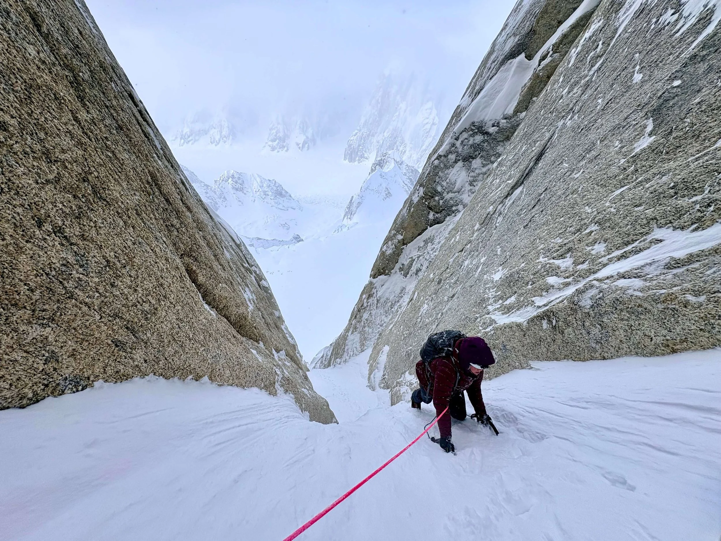 A woman in a dark red jacket is climbing a steep snow slope in a narrow couloir on the Ham and Eggs route. The mountains of the Ruth Gorge are visible through the clouds below.