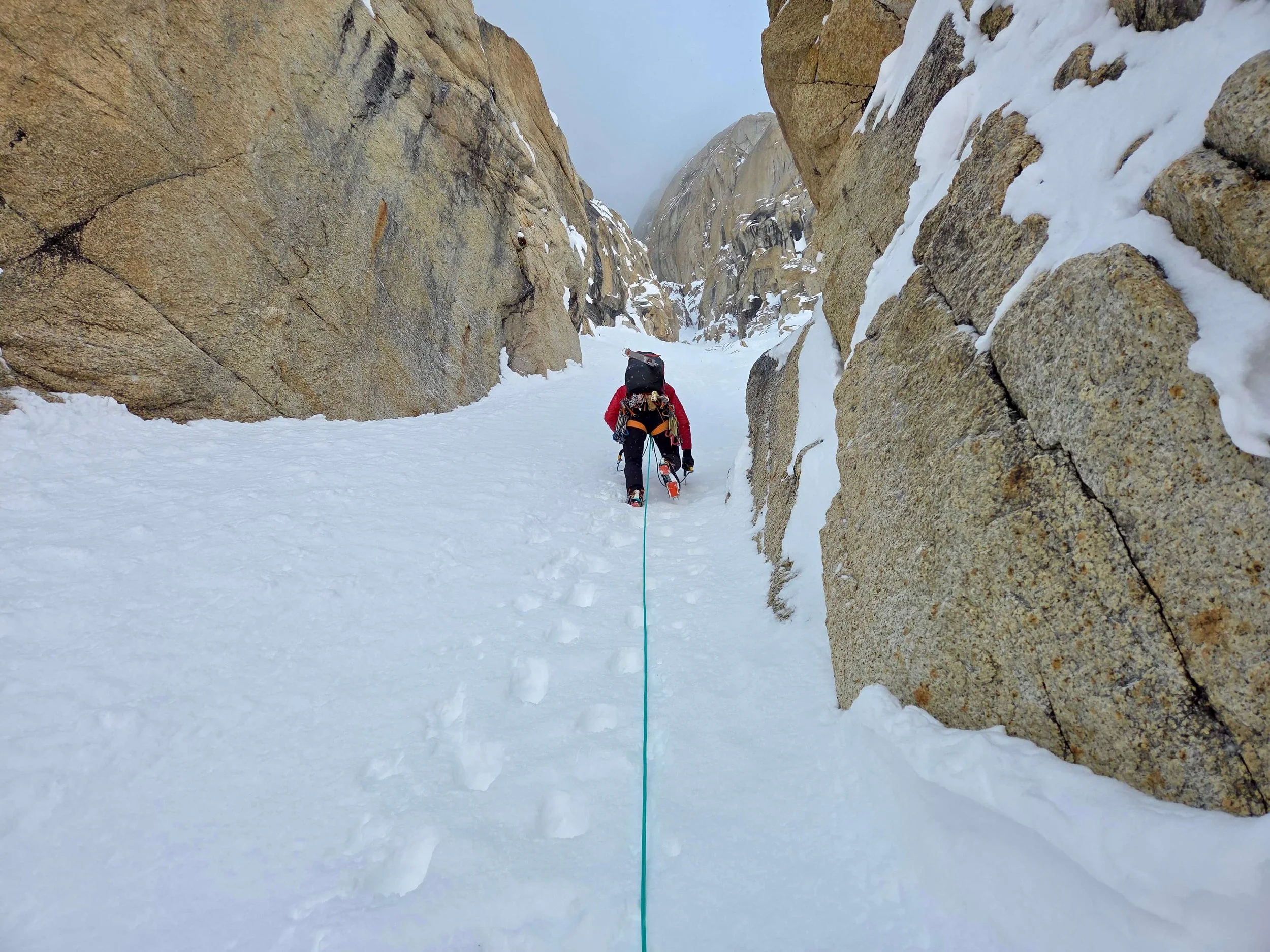 A Skyward Mountaineering guide is kicking steps up the steep snow at the start of the Ham and Eggs route on the Moose's Tooth. Steep walls of golden granite are on both sides of the couloir.