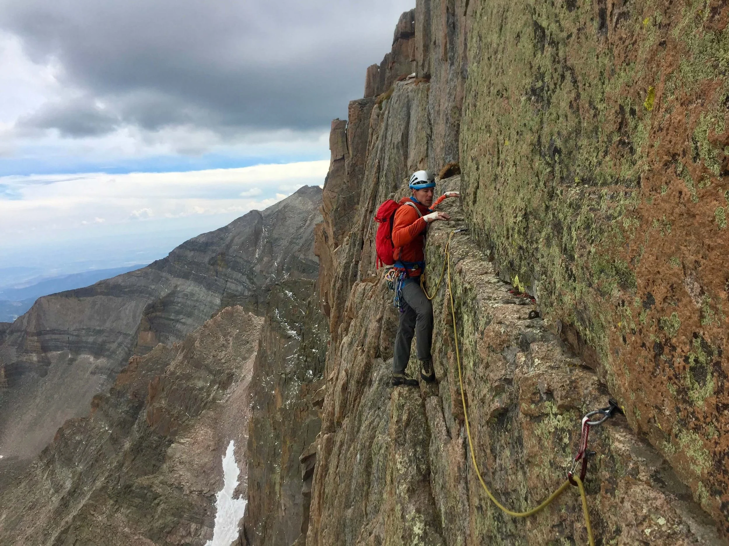 A guide is leading a traverse on the top pitch of the Casual Route on the Diamond of Long's Peak. He is wearing a blue helmet and red jacket.