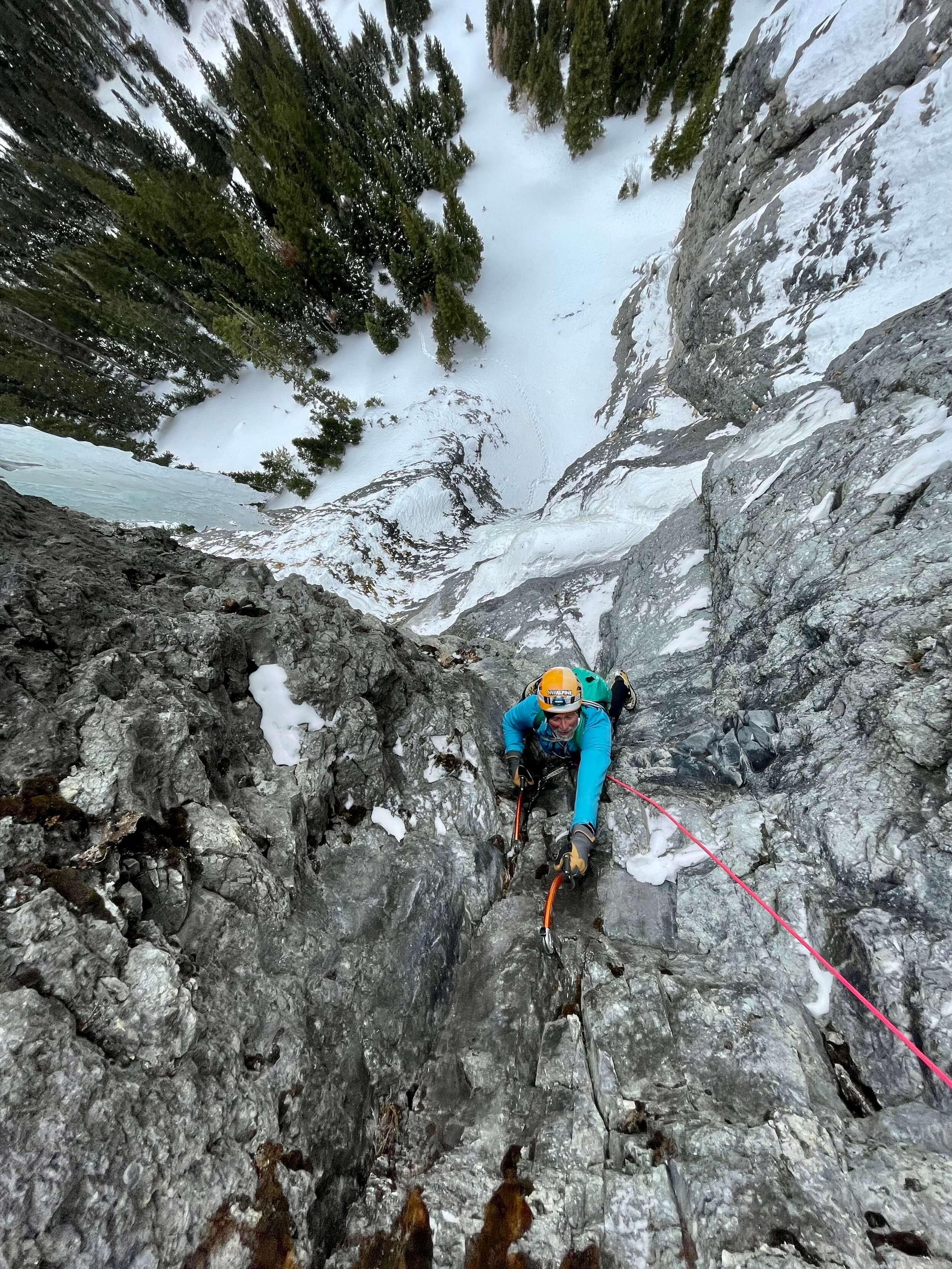 A person in a light blue jacket is mixed climbing a steep rocky corner with this ice tools and crampons. Pine trees and a steep snow slope are in the background below him.