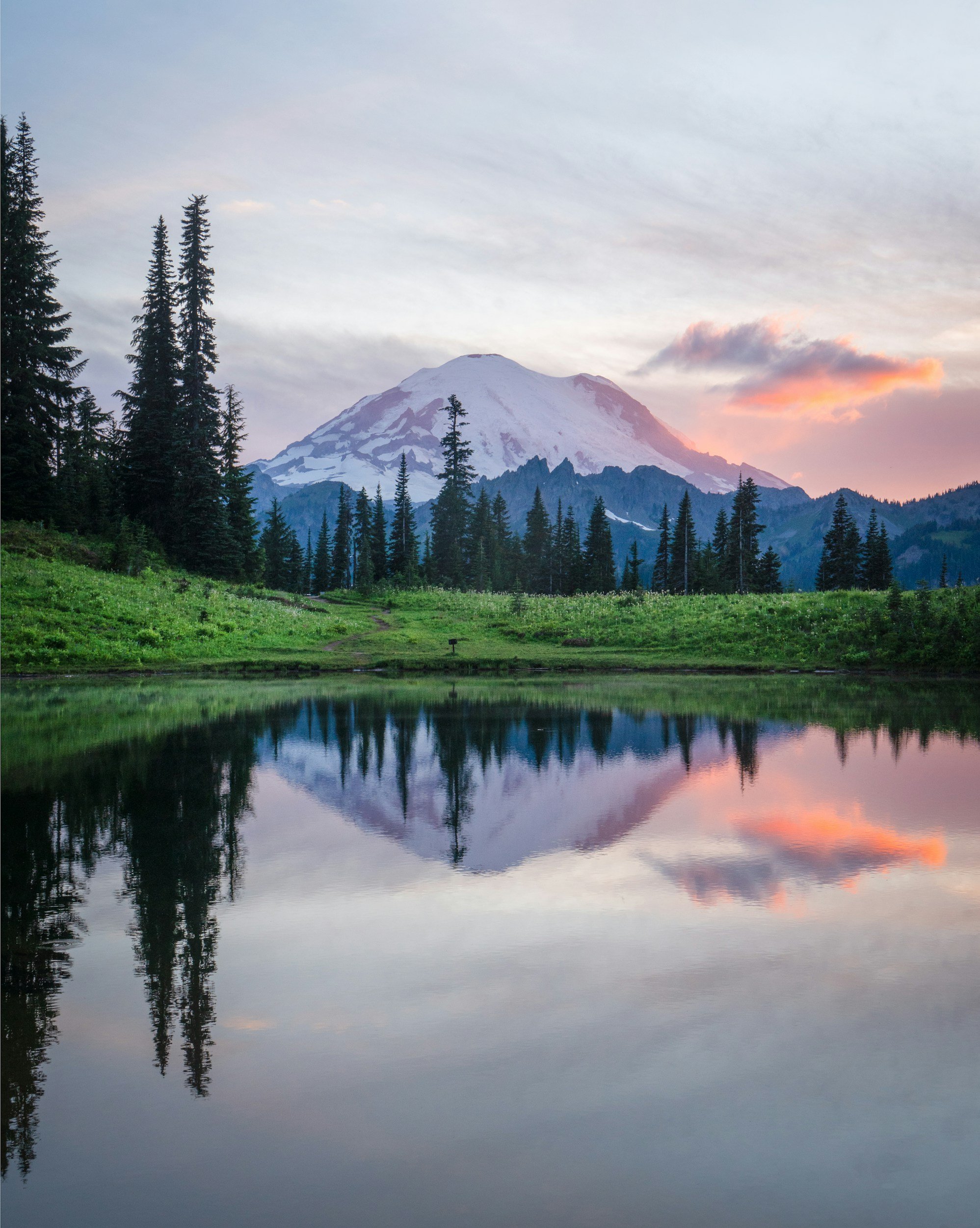 Snow-capped mountain behind pine trees, reflected in a calm lake during sunset.
