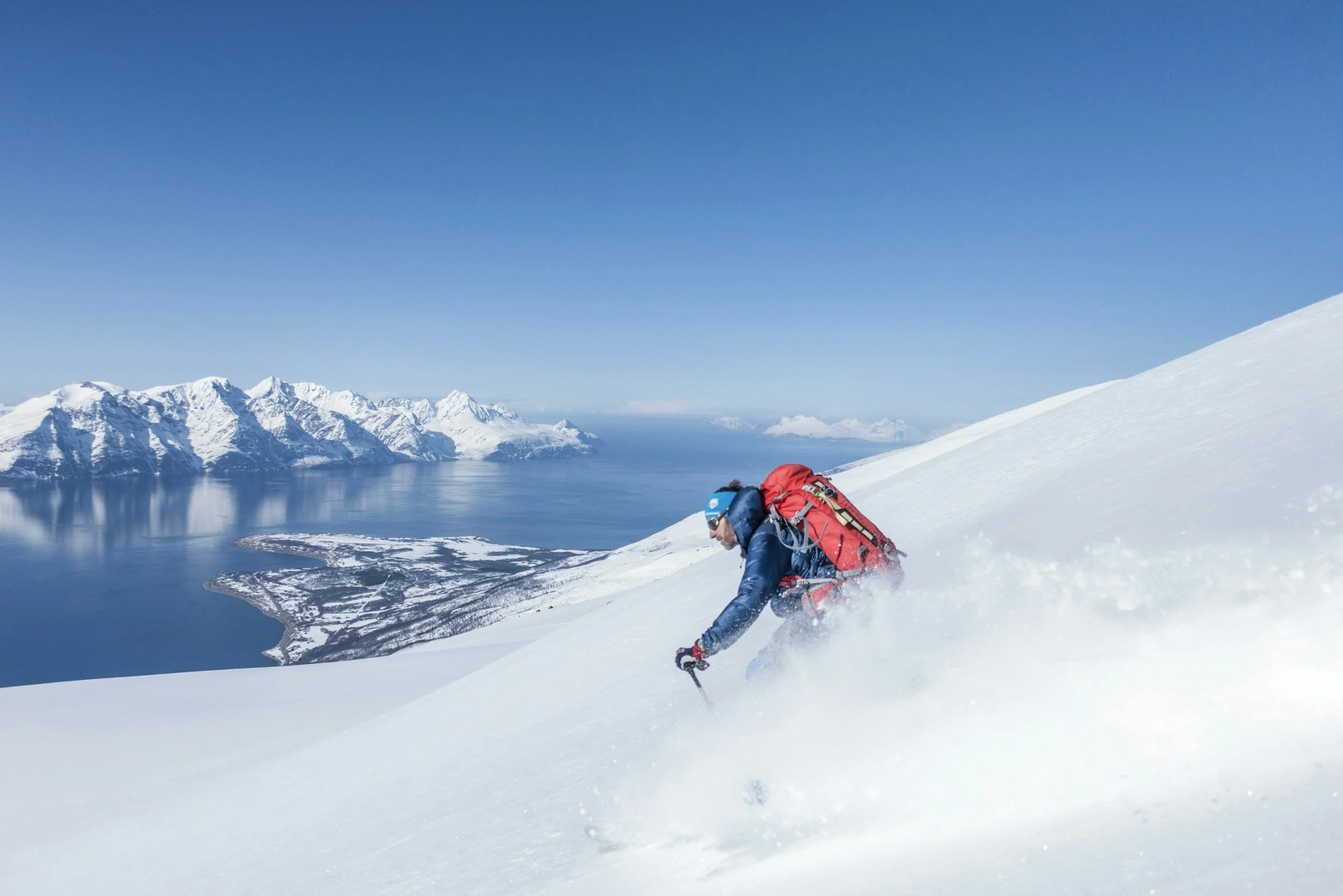A skier in a blue jacket and red backpack is skiing in deep powder in Greenland with an iceberg in the background.