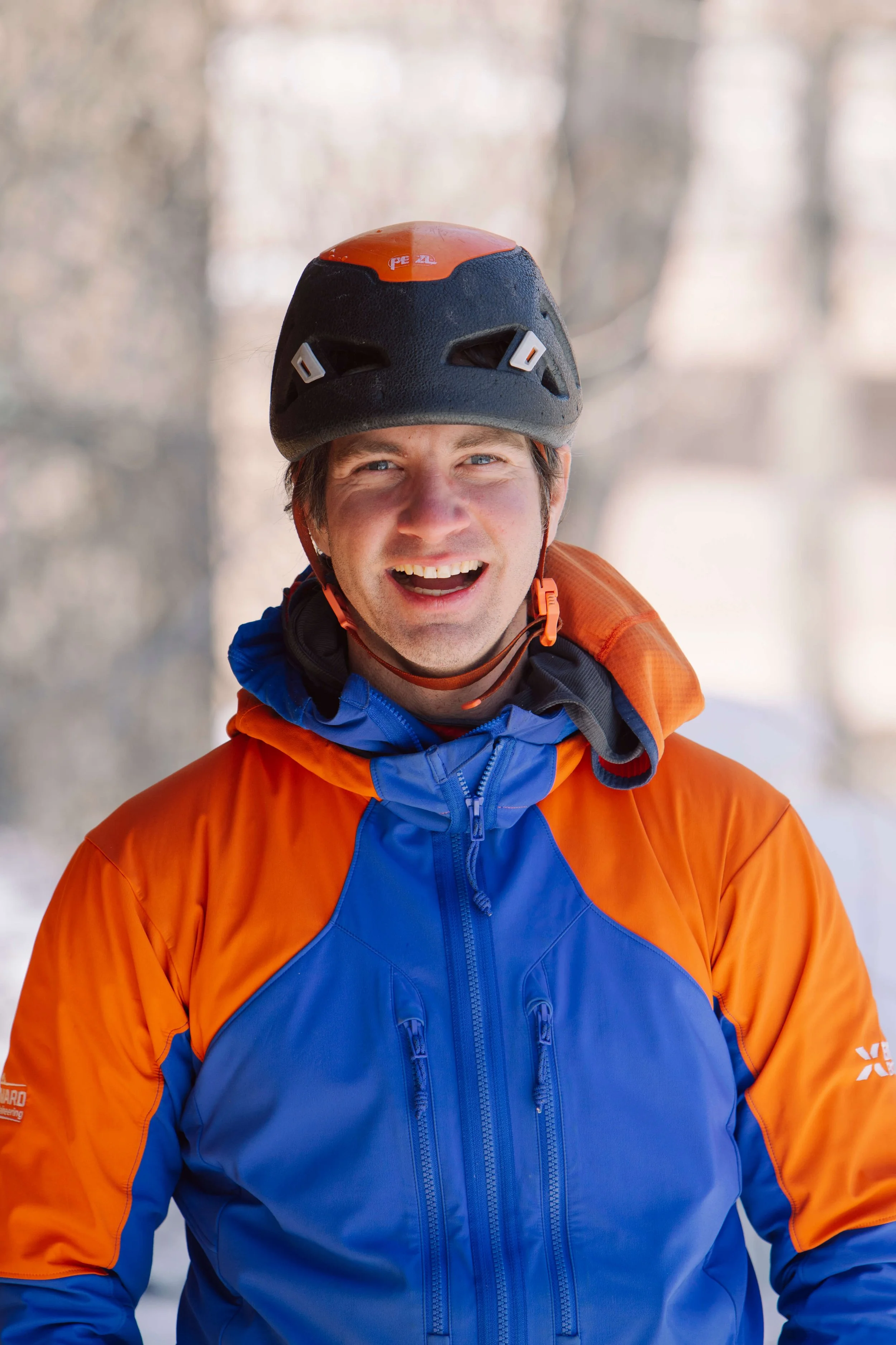 Skyward Mountaineering guide Bobby Cosker is smiling during a day of guiding ice climbing in the Ouray Ice Park. He is wearing a black helmet and orange and blue jacket..