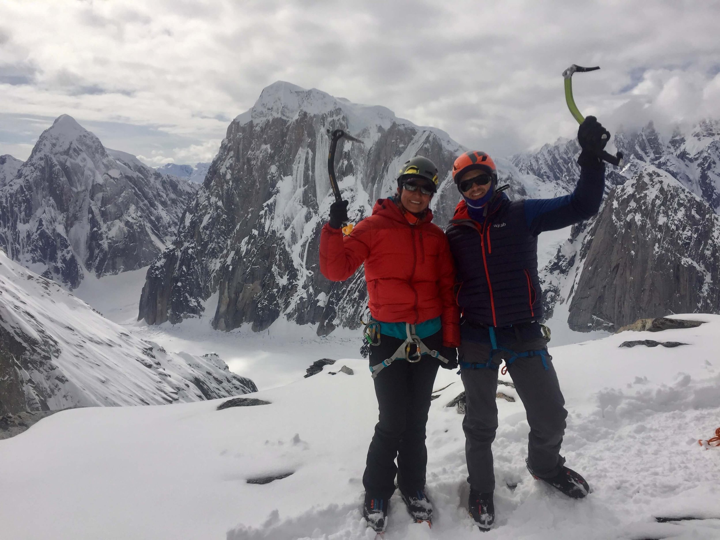 Two climbers, one in a red jacket and the other in a blue jacket, stand on the summit of the Wisdom Tooth above the Ruth Gorge. They are each holding an ice tool above their heads in celebration.