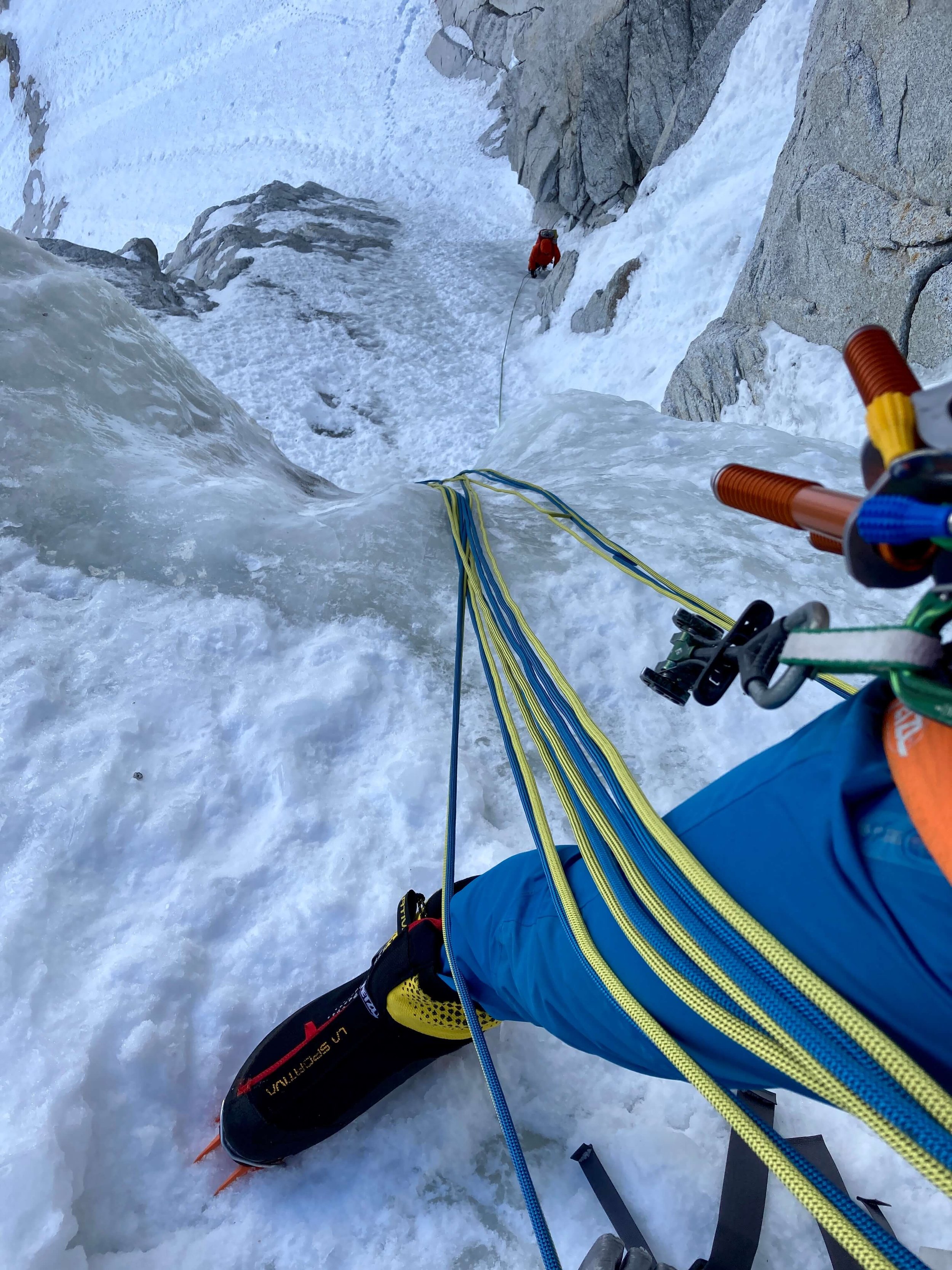 A blue and yellow rope is draped over the belayer's leg, wearing blue pants with ice screws and cams hanging off the harness. The climber below is starting up a pitch of steep snow and ice.