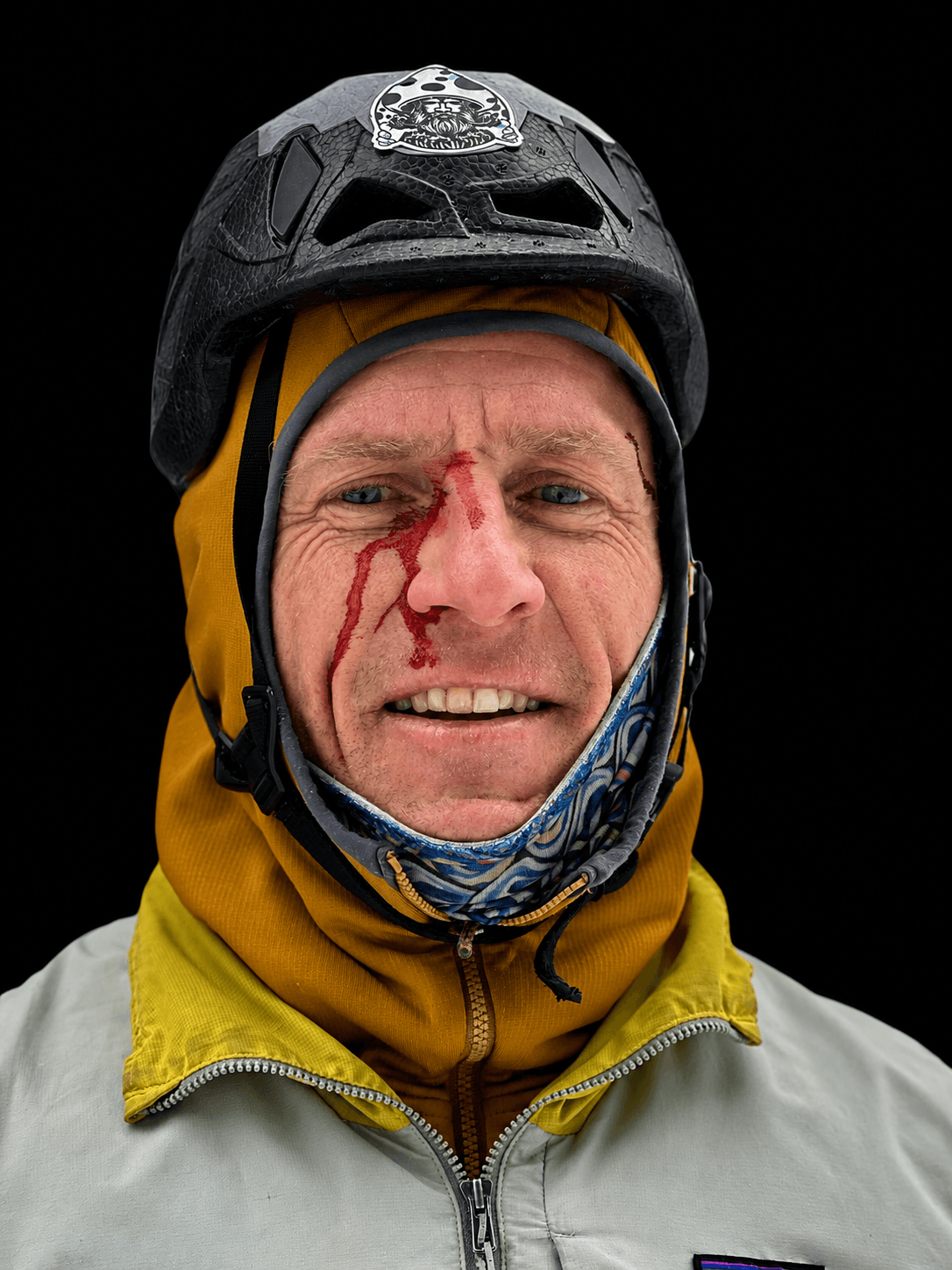 Skyward Mountaineering founder and IFMGA Guide, Vince Anderson is smiling during a day of instructing in the Ouray Ice Park. He is wearing a yellow jacket with a black helmet and has blood on the bidge of his nose and cheek from getting hit by ice.