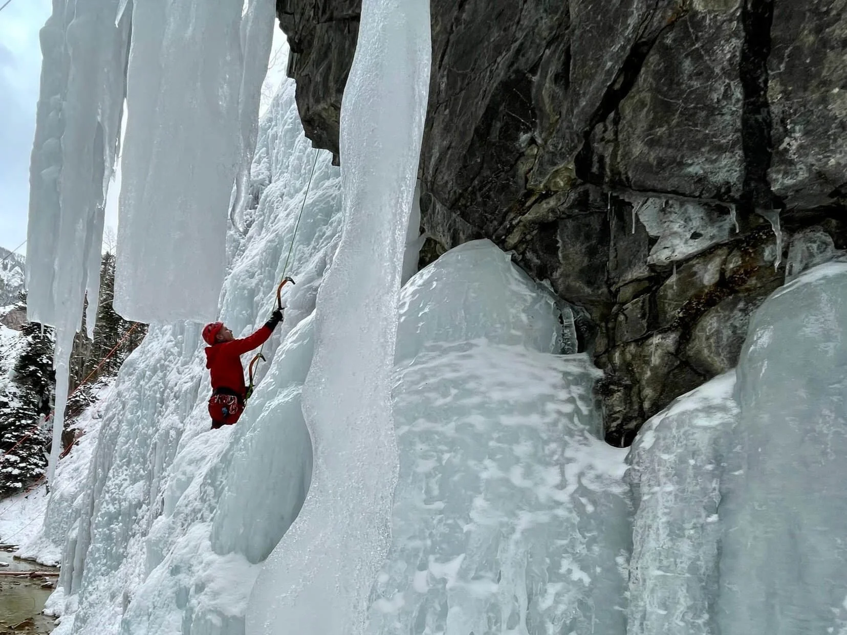 A person in a red jacket is ice climbing in the background amidst dripping icicles inside a rocky cave.