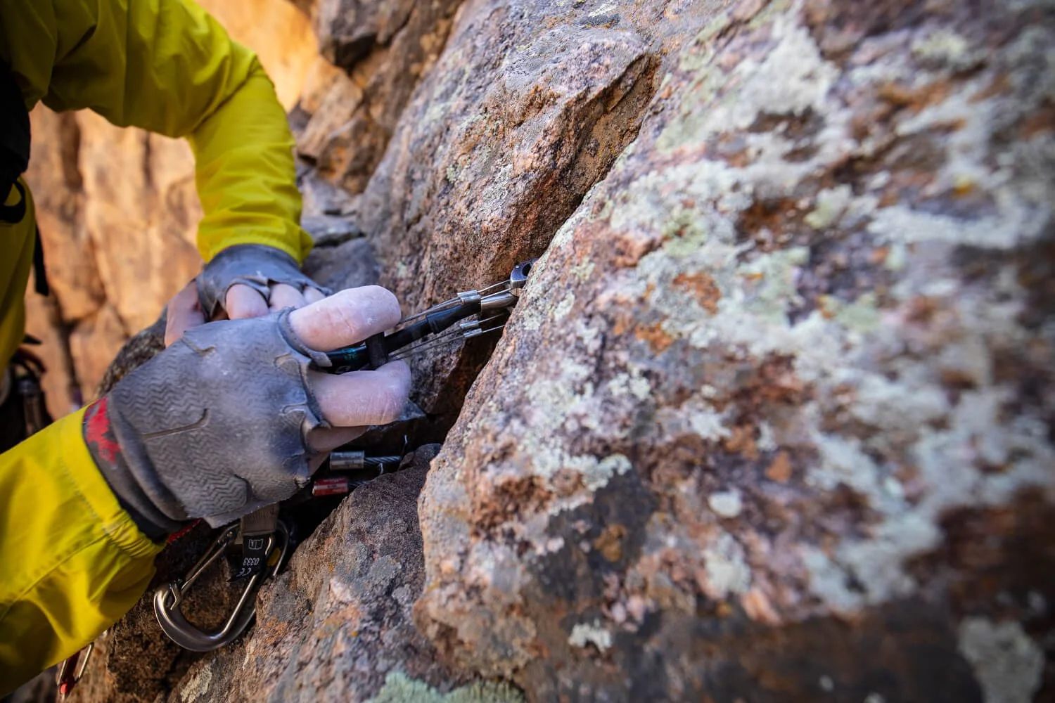 A guide in a yellow jacket and black crack climbing gloves is placing a piece of climbing protection in the crack to build an anchor.