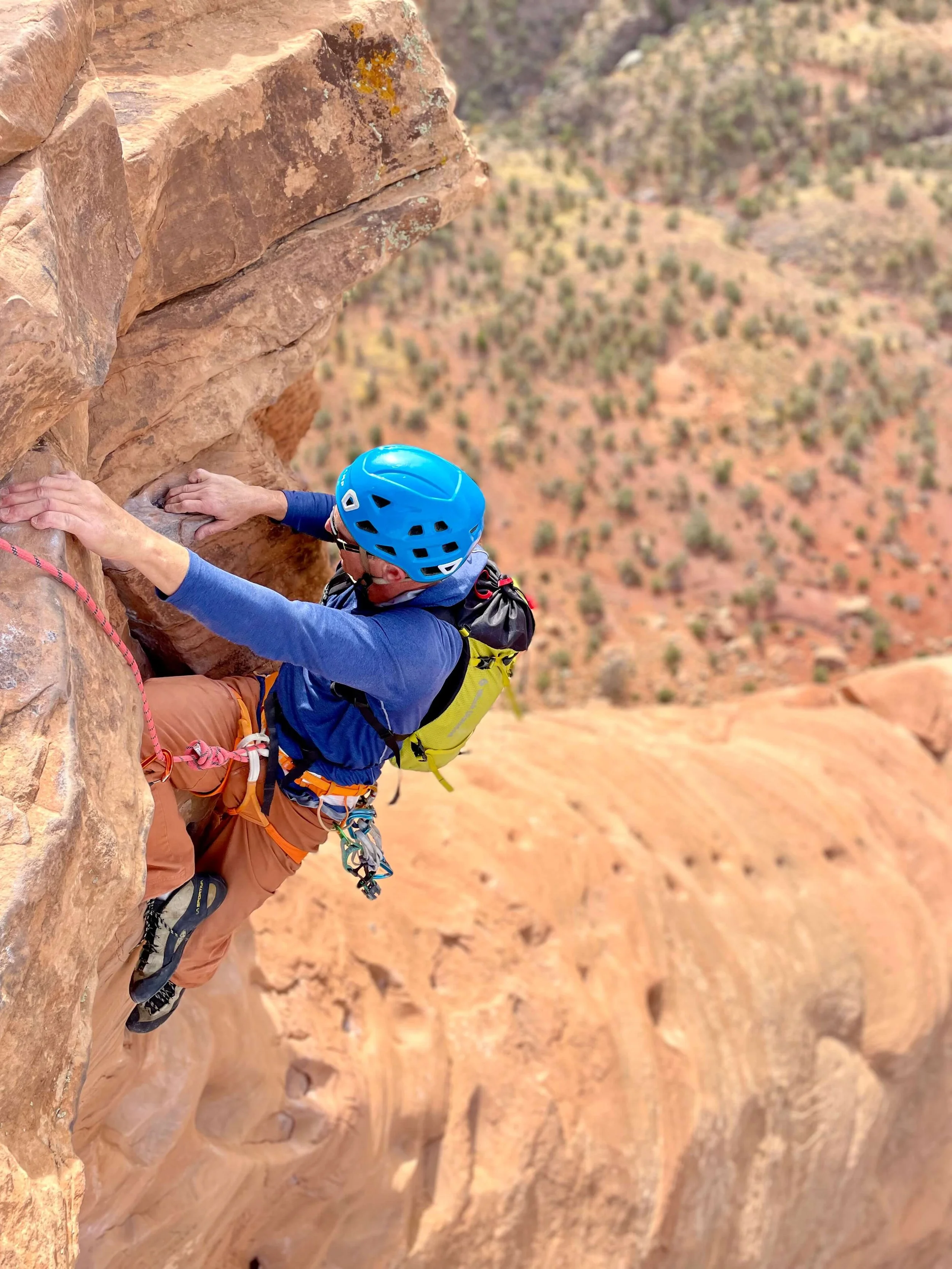 A rock climber in a yellow backpack and blue helmet is climbing the steep headwall on Otto's Route in Colorado National Monument.
