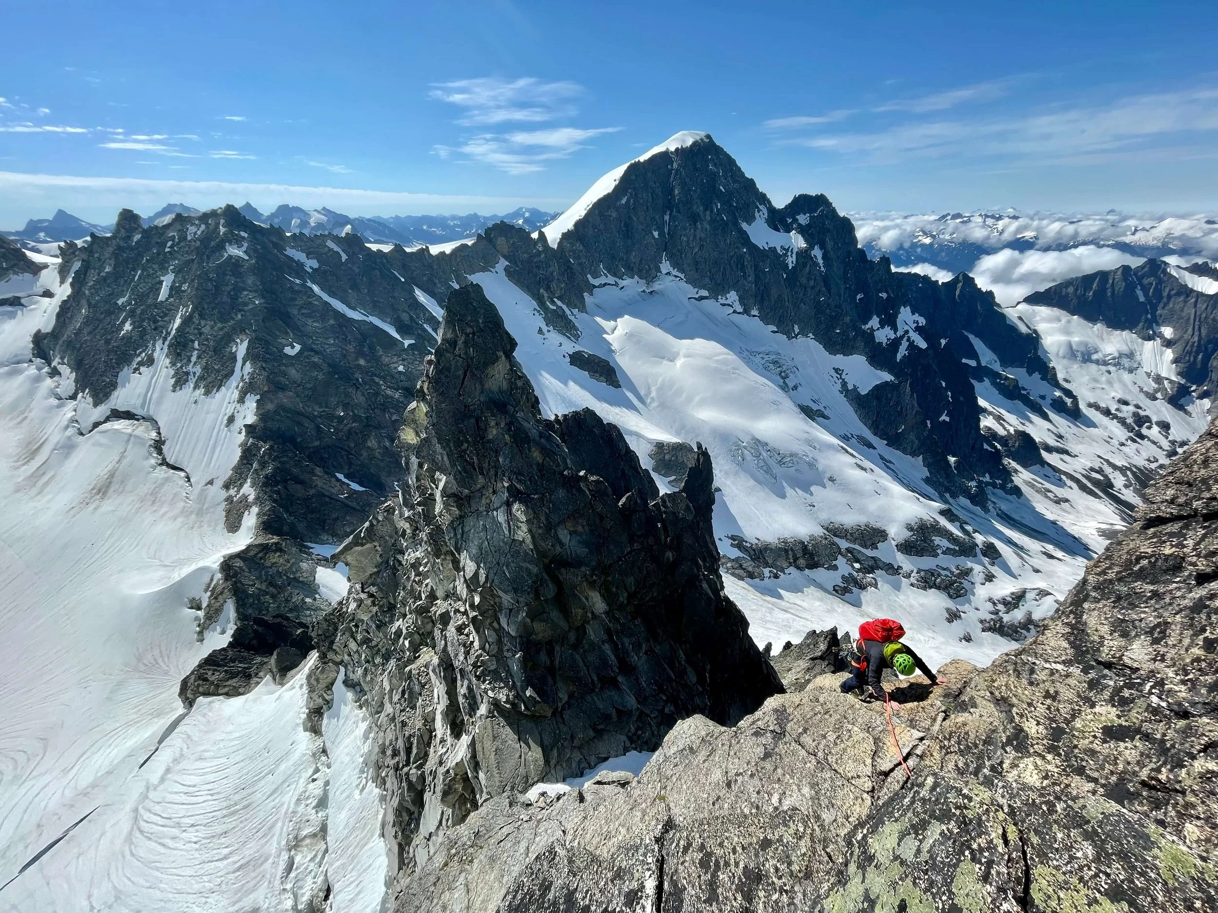 A climber in a green helmet and red backpack is ascending the Northeast Ridge on Dorado Needle with the summit of Eldorado Peak in the background.