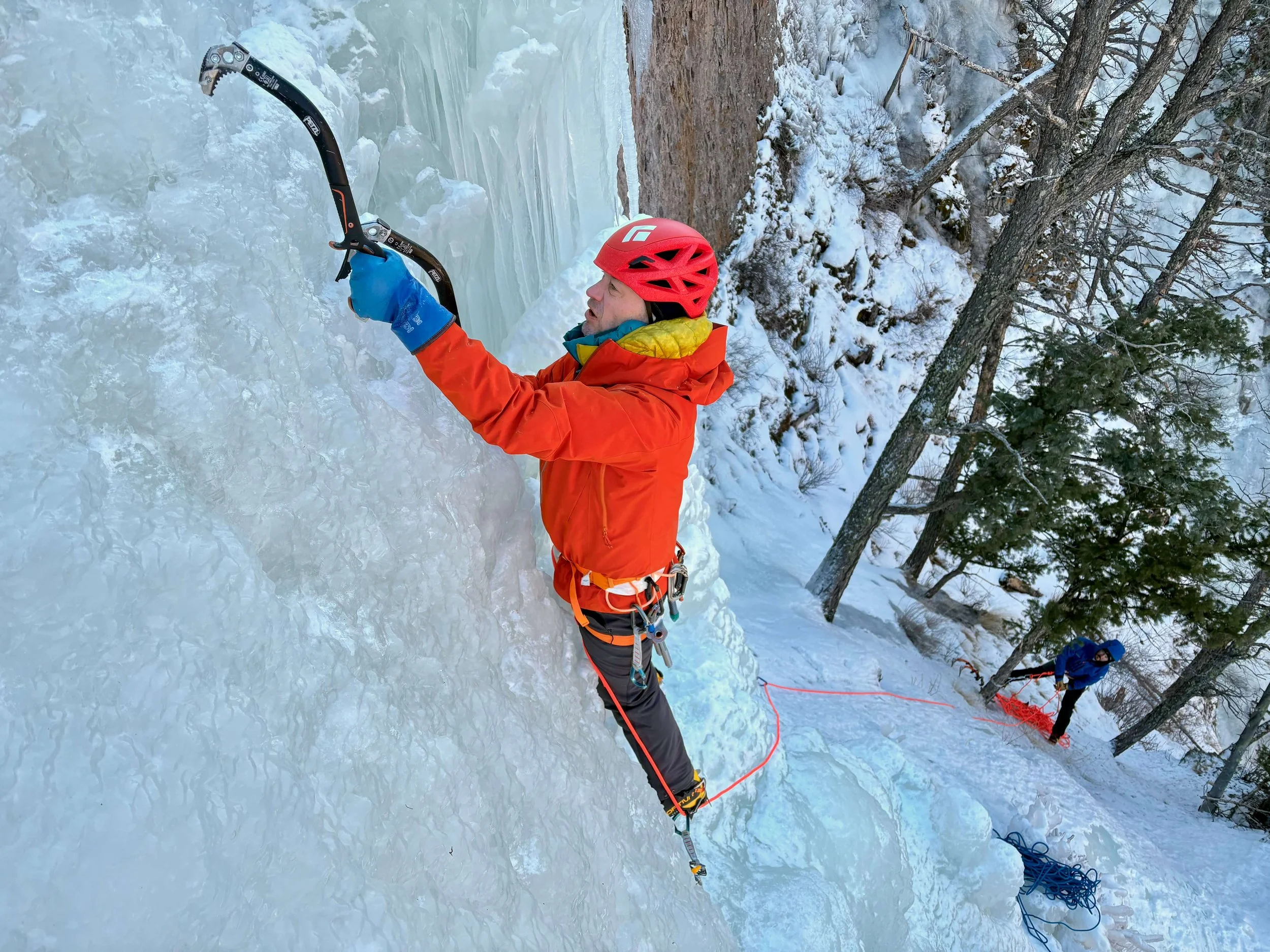 An ice climber in an orange jacket and red helmet is a leading a multipitch route in the Lake City Park.