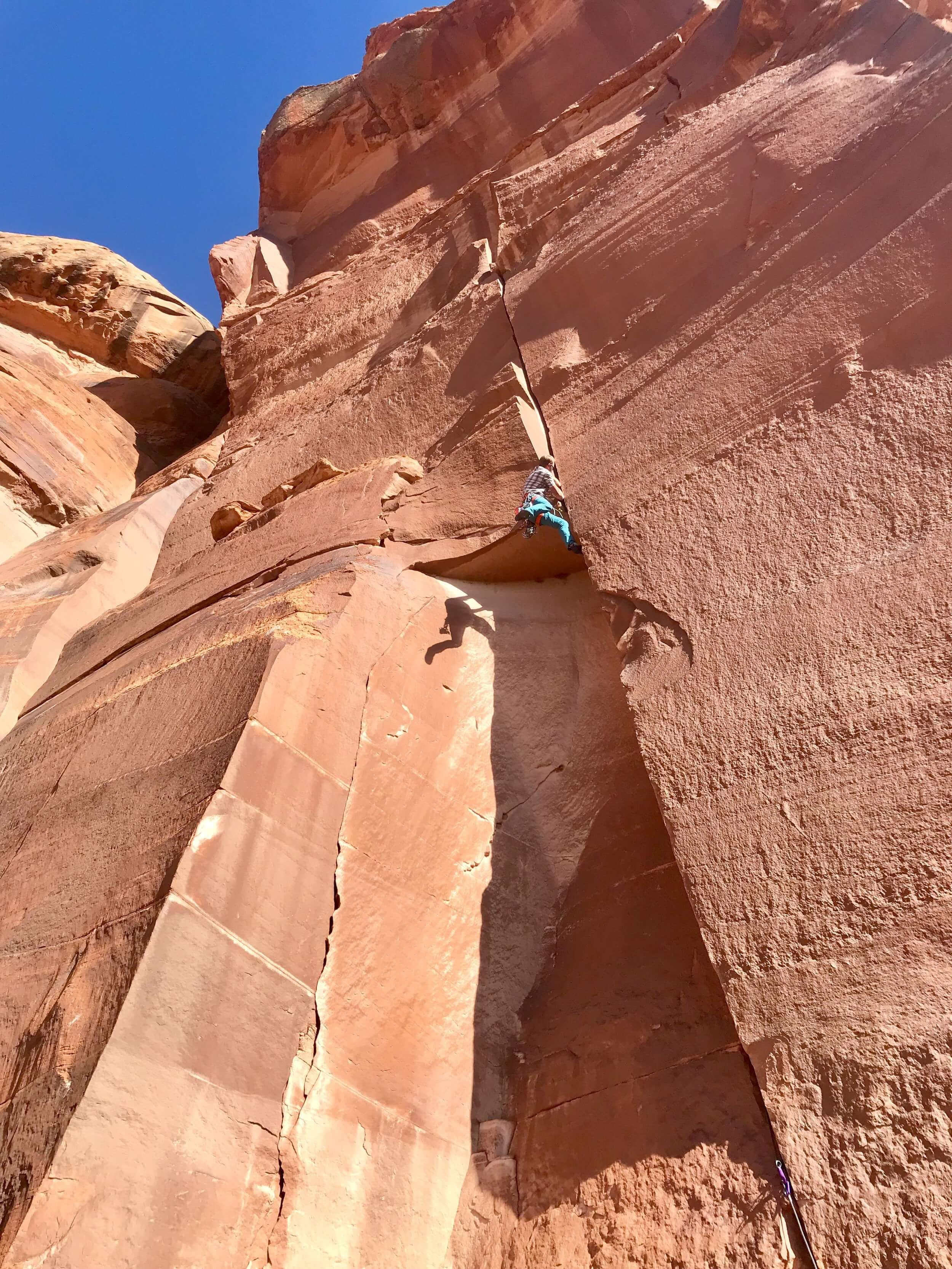 A person rock climbing on a tall red sandstone cliff under a clear blue sky. He is climbing past a small roof feature in the middle of the cliff.