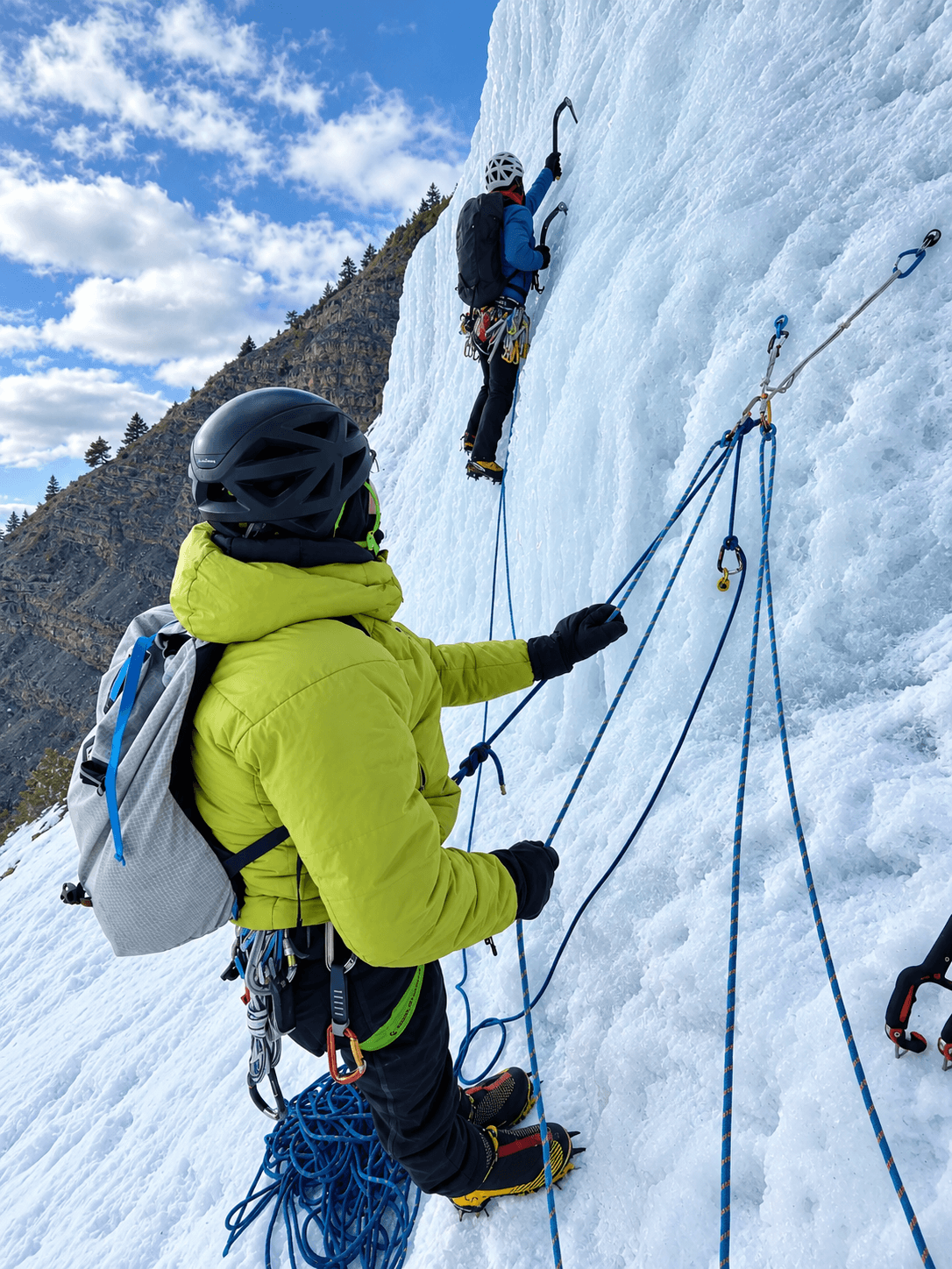 A climber in a yellow jacket is belaying the lead climber on the route, Dexter Creek Slabs during a Lead Ice Climbing Course in Ouray, Colorado.