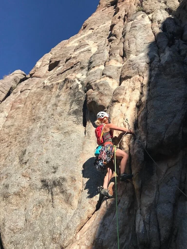 A woman rock climbing on a steep cliff face, wearing a white helmet is leading a pitch on golden granite in Unaweep Canyon.
