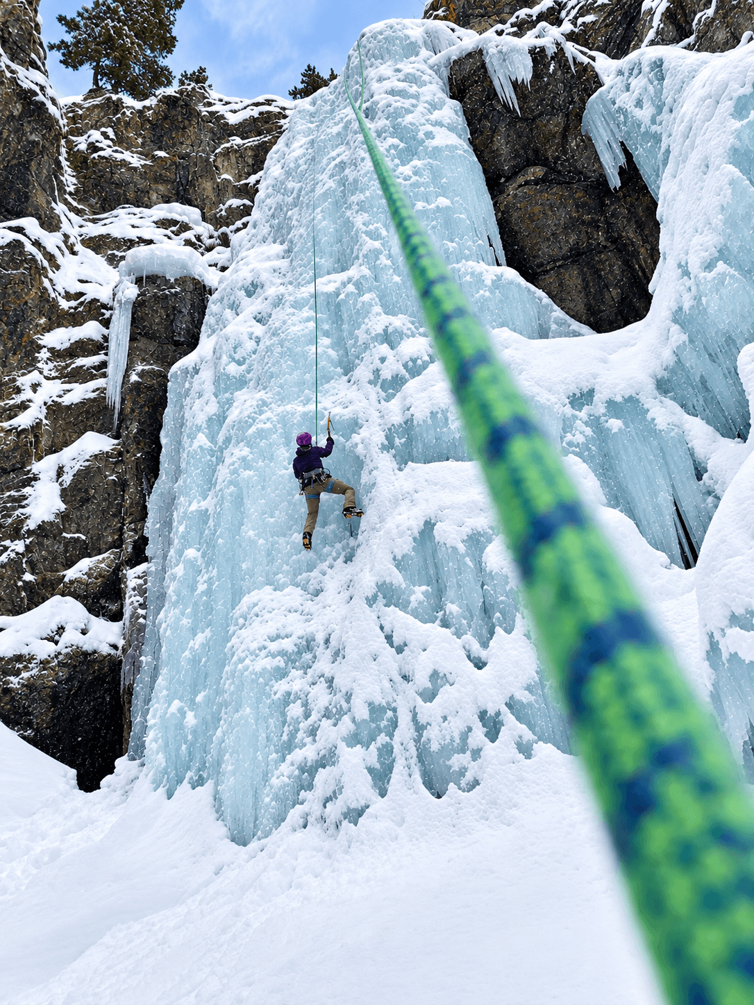 A climber is toproping an ice climbing route on Camp Bird Road during an alpine climbing course. A green rope is in the foreground being belayed by the guide.