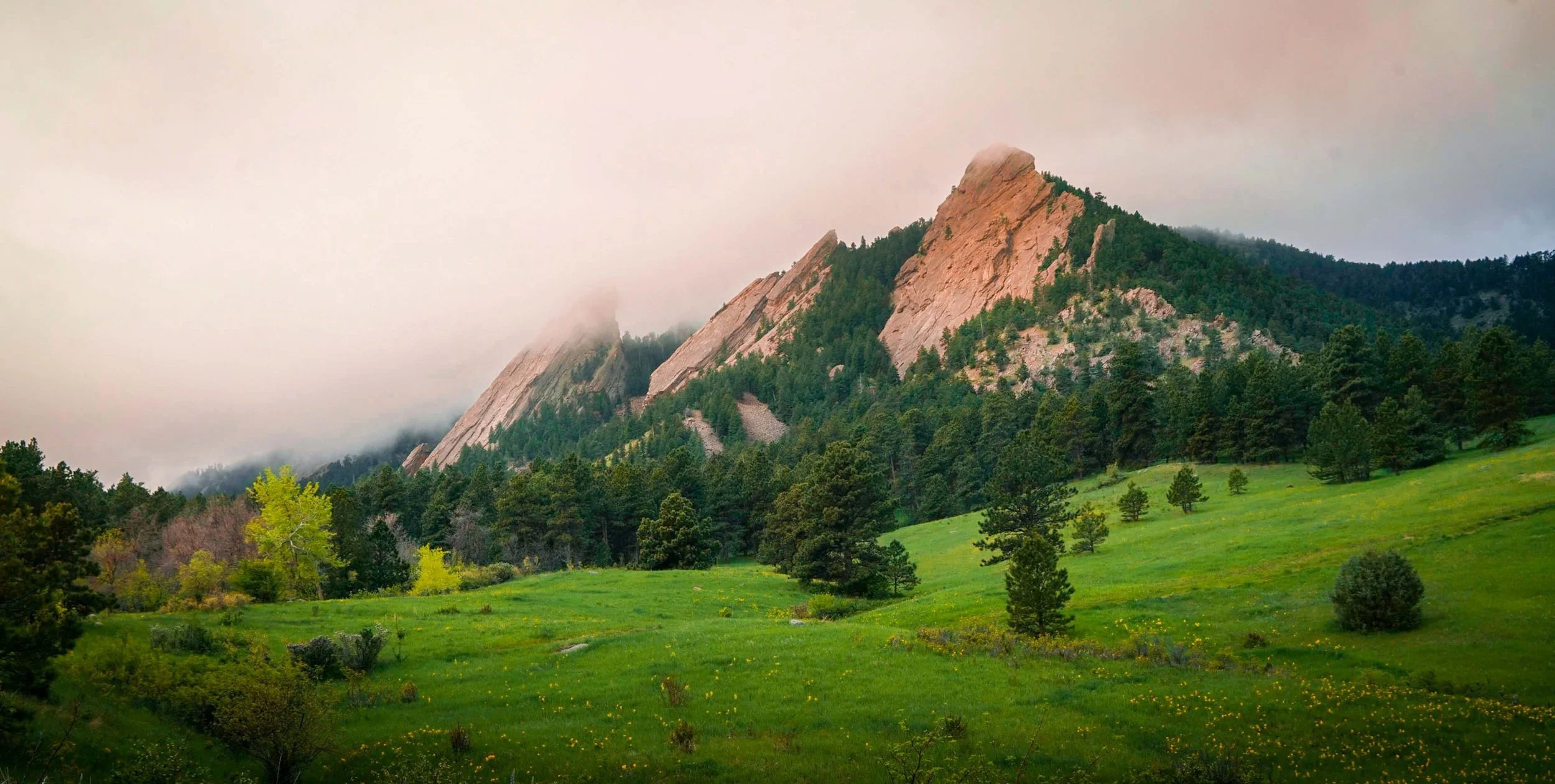 Clouds roll over the cliffs on Boulder's Flatirons with lush green grass in the foreground.