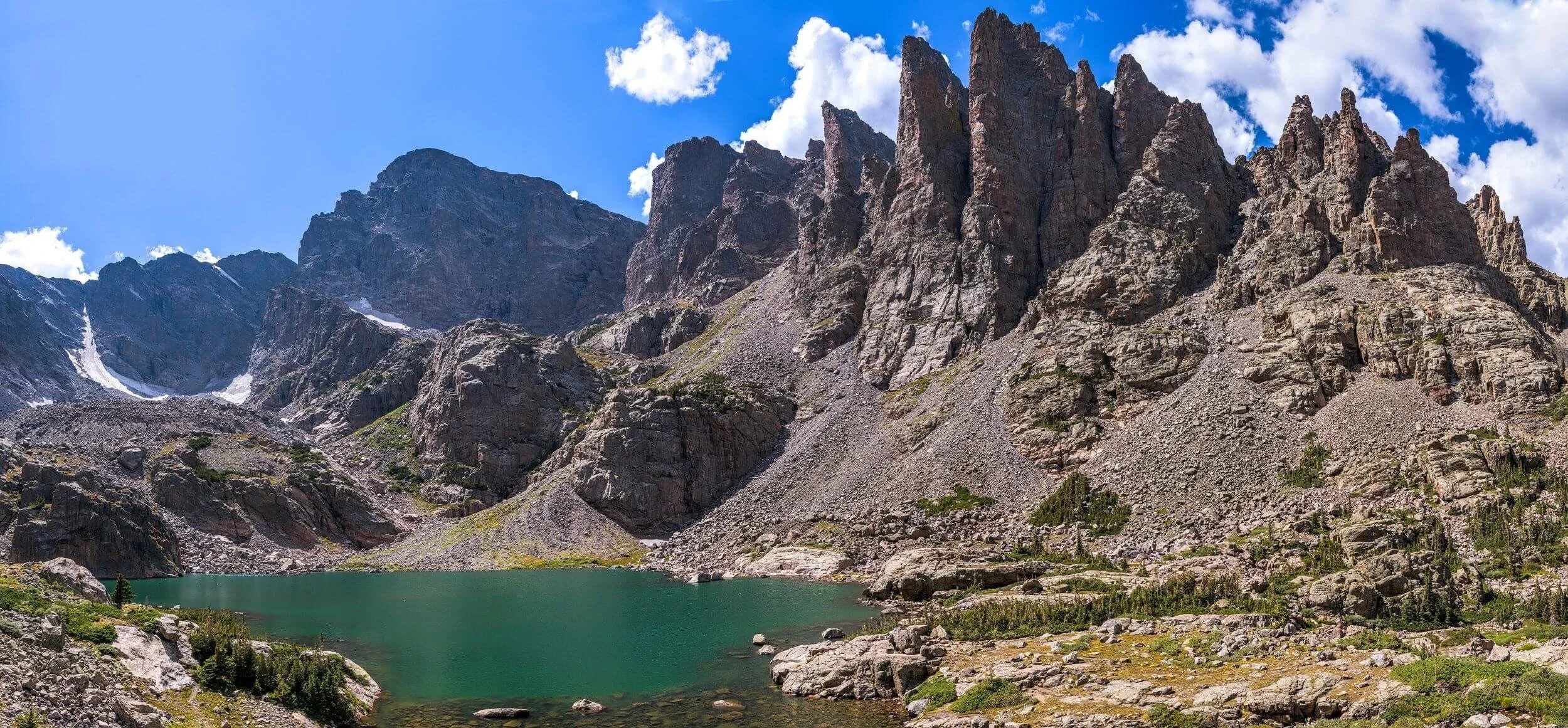 Mountain landscape of Sky Pond and the Petit Grepon with a turquoise alpine lake, rocky peaks, and scattered greenery under a blue sky with clouds.