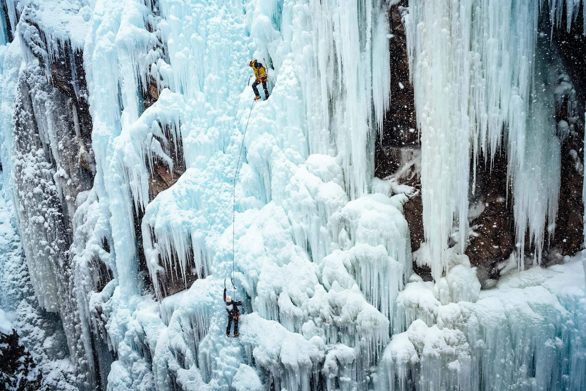 A woman in a blue jacket is ice climbing in the Ouray Ice Park, being belayed from above by a climber in a yellow jacket.