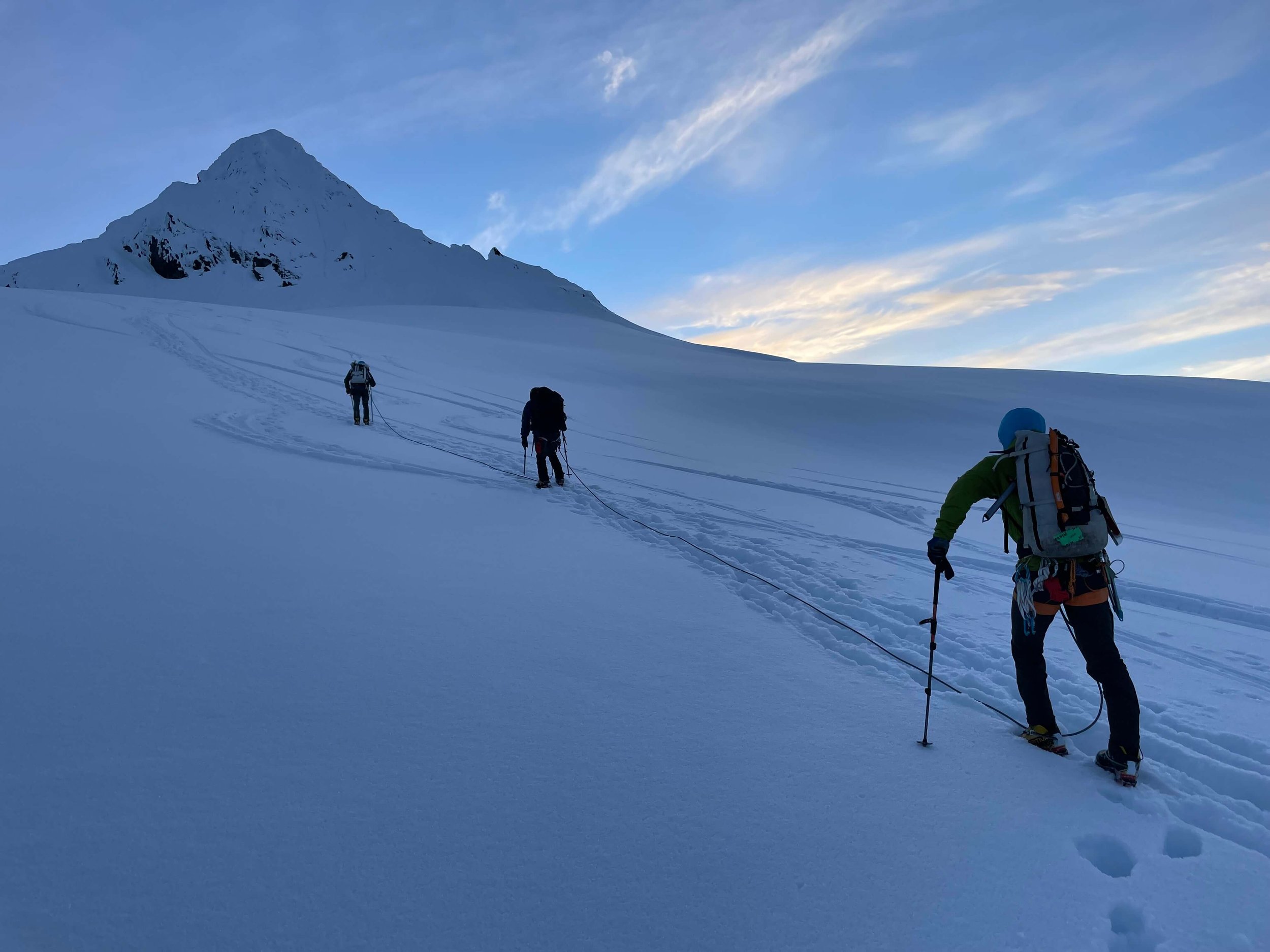 A rope team of three mountaineers are climbing up a snowy glacier towards Mount Shuksan's summit pyramid. The sun is starting to rise from behind the peak casting an alpenglow shadow.