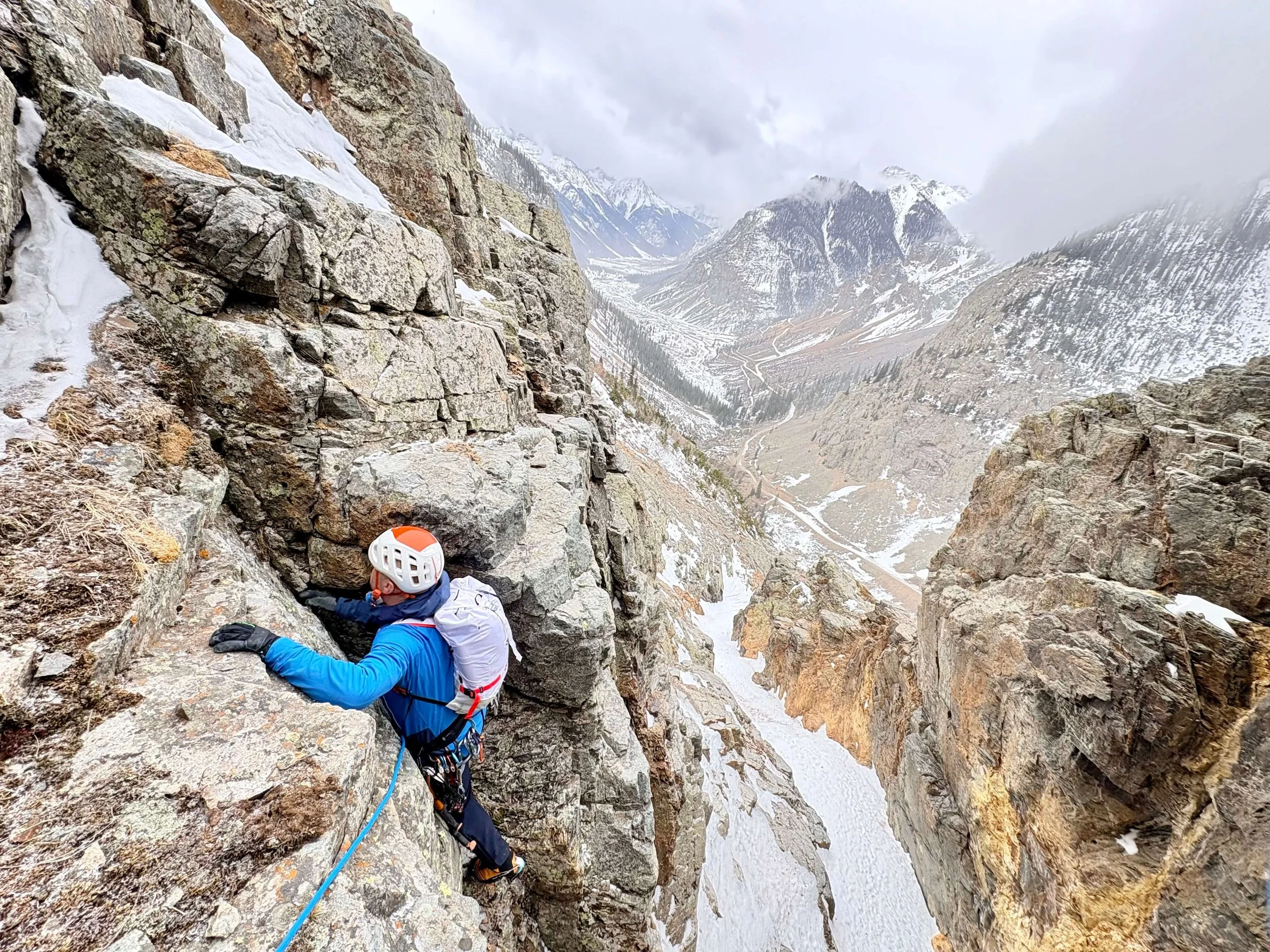 A person in a blue jacket and white helmet is climbing a mixed pitch on Hoser's Highway in Eureka, Colorado with mountains in Silverton in the background.