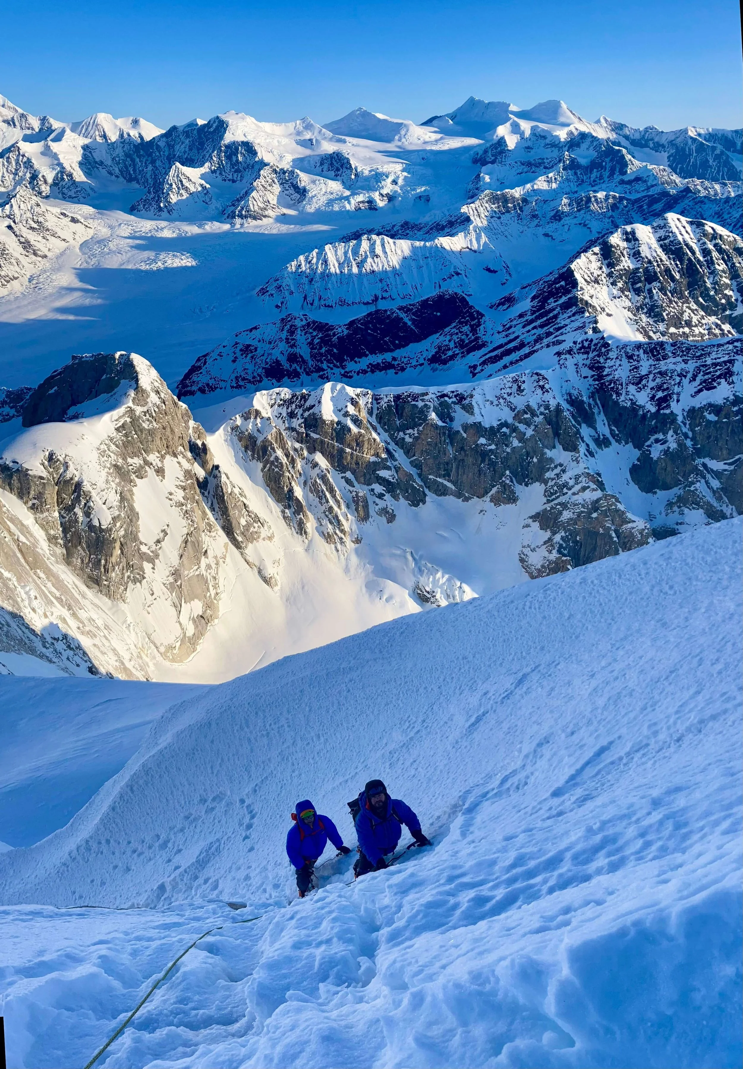 Two climbers are ascending a steep snow slope to the summit of the Moose's Tooth with sun lighting up the mountains of the Ruth Gorge glacier in the background.