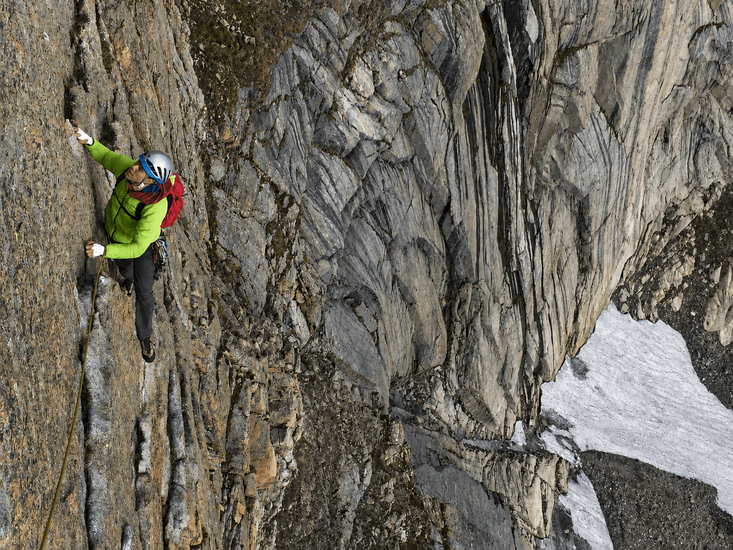 A rock climber in a lime green jacket, blue helmet and red backpack is climbing a large granite face.