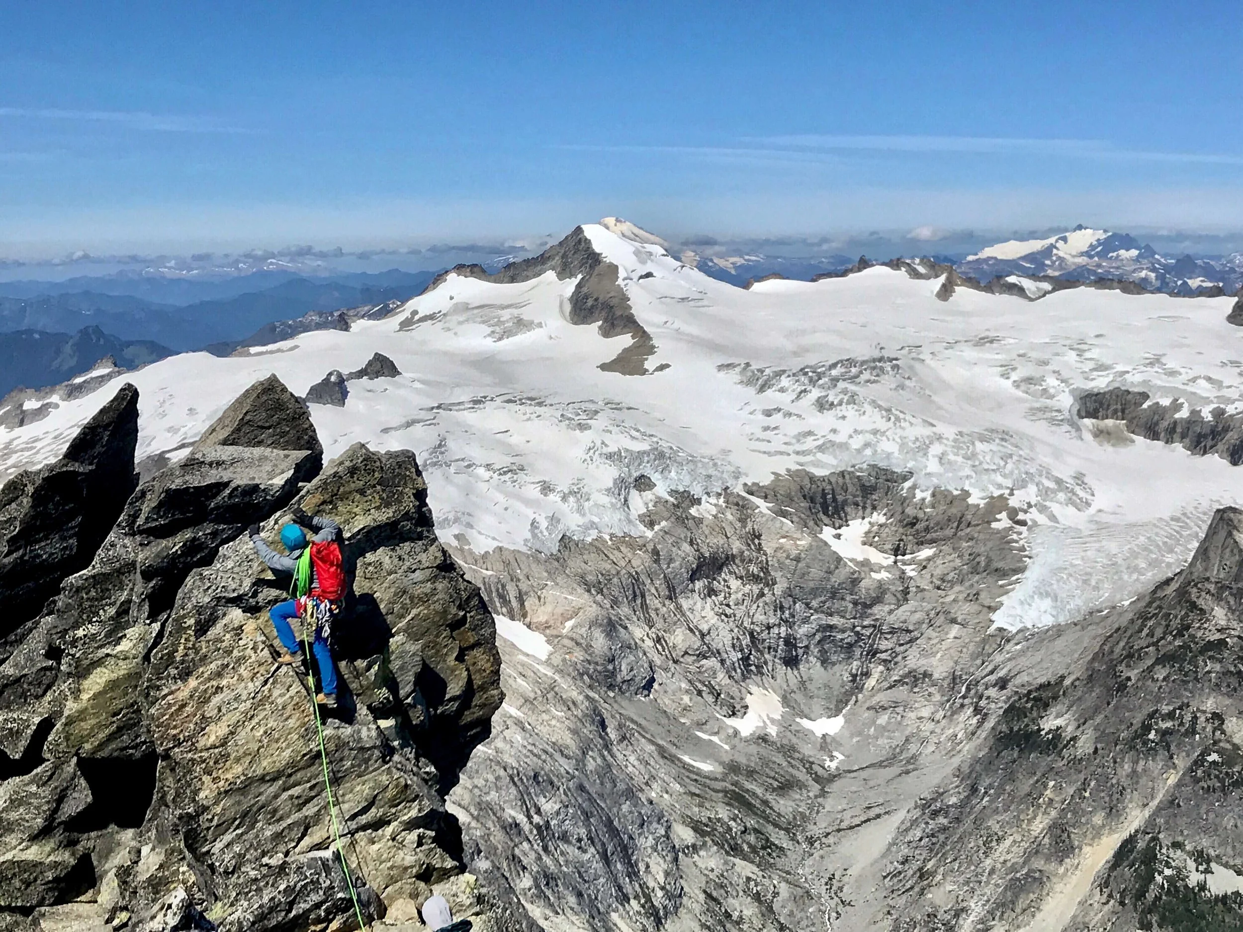 A guide wearing a red backpack leads up the summit block on the West Ridge of Forbidden Peak with the glaciated Eldorado Peak in the background. 