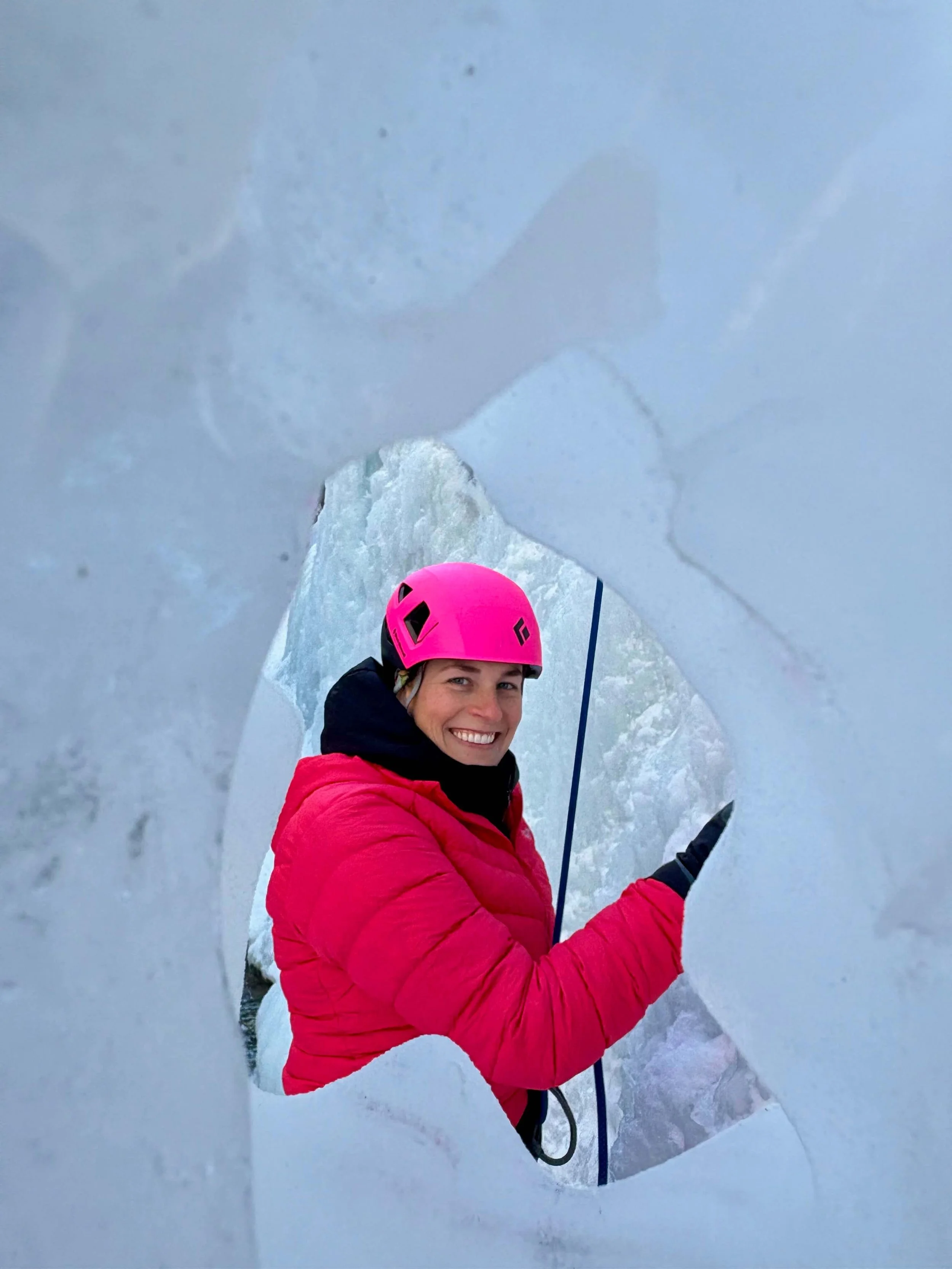 A climber in a pink helmet and pink jacket is belaying, looking through an icicle in the Ouray Ice Park.